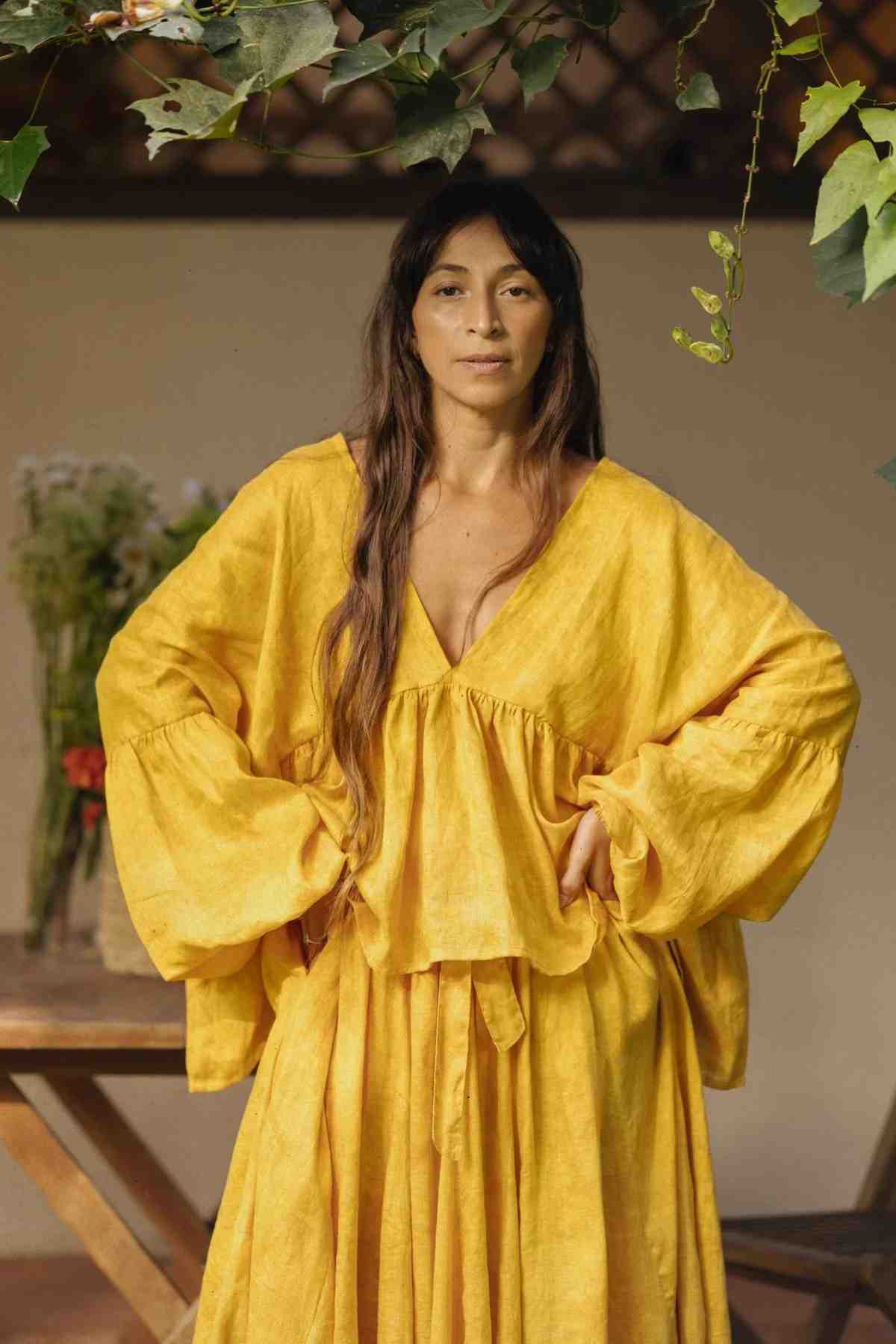 A person with long brown hair stands confidently with hands on hips, wearing a loose-fitting, brightly colored Linen Eva Top & Orisa Dance Skirt Set from Myrah Penaloza. They are against a blurred background featuring green foliage and a wooden table with a flower arrangement.