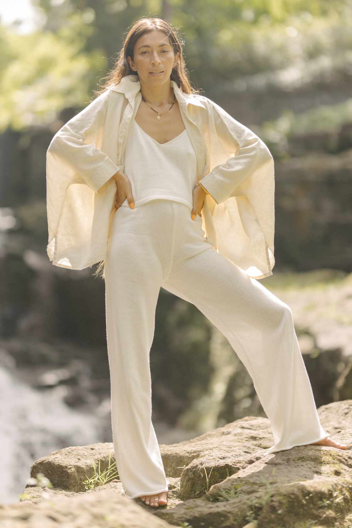 A woman stands confidently on a rock in an outdoor setting, wearing a flowing white outfit that features the everyday staple, the Grace Jacket by Myrah Penaloza (100% Linen). With her hands on her hips and long brown hair cascading down, she is surrounded by a blurred natural background of greenery and sunlight.