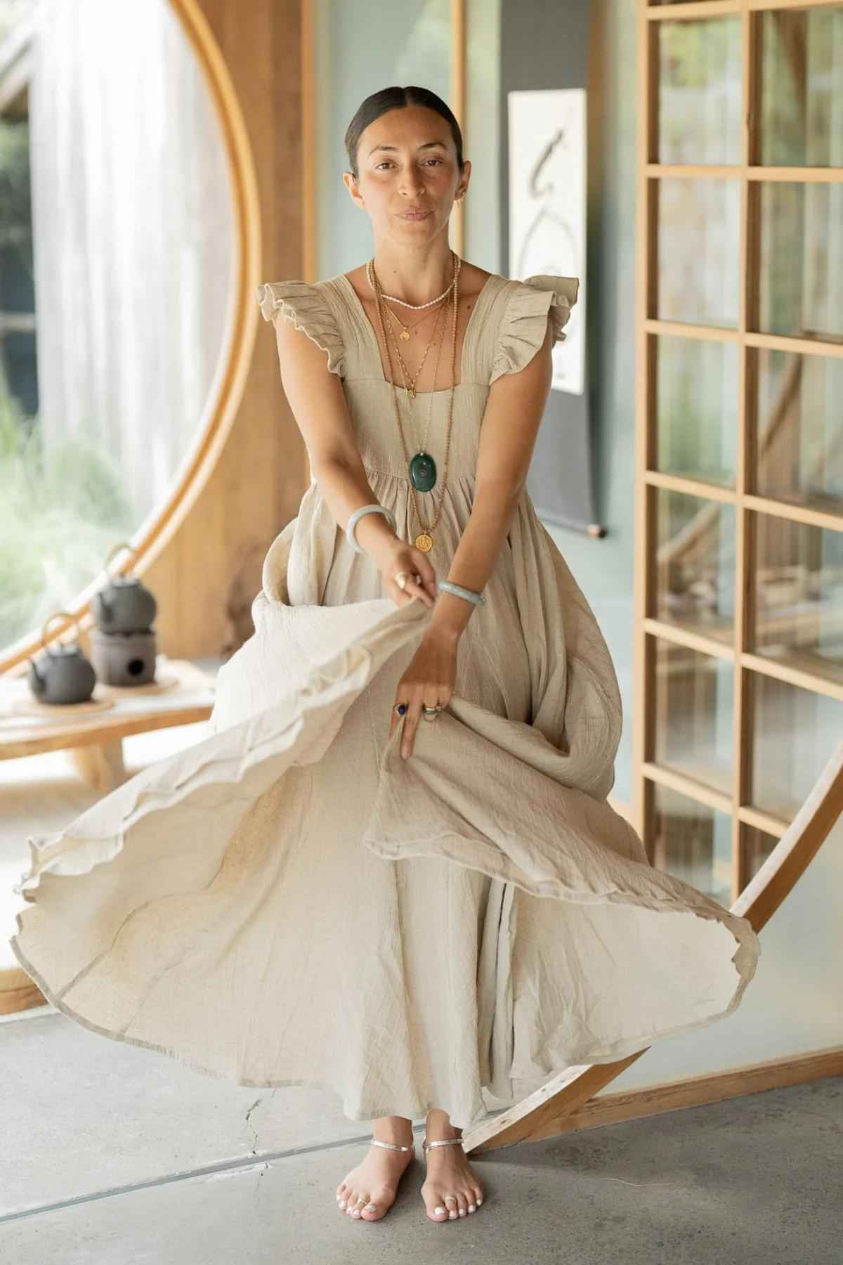 A person stands barefoot on a stone floor, adorned in a flowing, light beige Linen Anna Yemaya Gown with ruffled sleeves from Myrah Penaloza. They are accessorized with several necklaces and bracelets, and their hair is pulled back. The background showcases a circular window and wooden grid-style partitions.