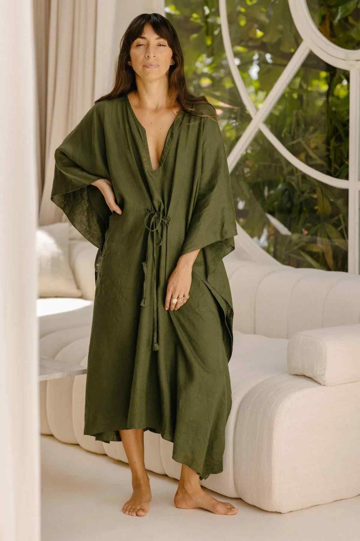 A woman stands barefoot on a light-colored floor in a bright room with large windows and lush green plants visible outside. She is wearing a long, loose-fitting River Kaftan by Myrah Penaloza, made of olive-green stone-washed linen, featuring a deep V-neck and drawstring waist. Her long dark hair cascades down her back as she appears relaxed and serene.