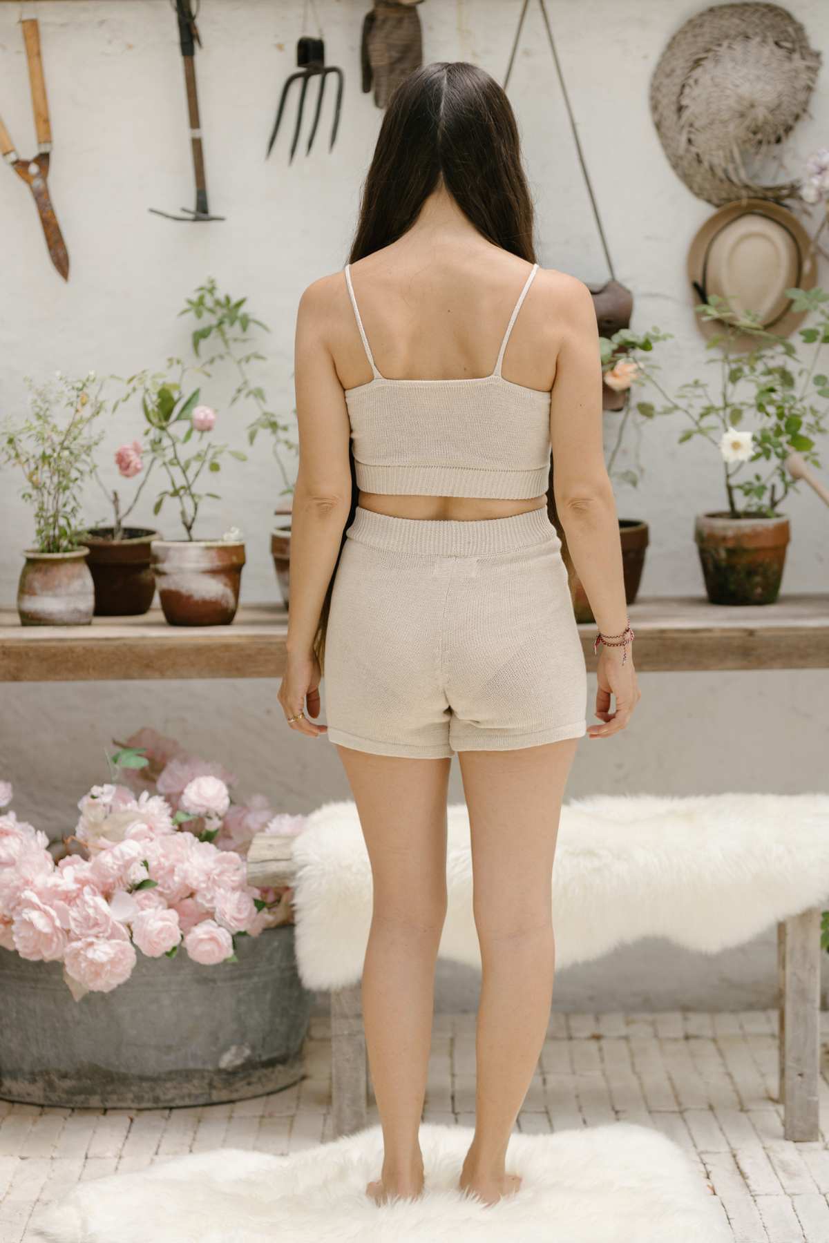 A person with long hair is standing in front of a wooden table adorned with potted plants and gardening tools on the wall. They are wearing a beige tank top and shorts from the sustainable Honey Knit Sets collection by Myrah Penaloza. Nearby, pink flowers bloom in a tub, while wool rugs are draped over the bench.