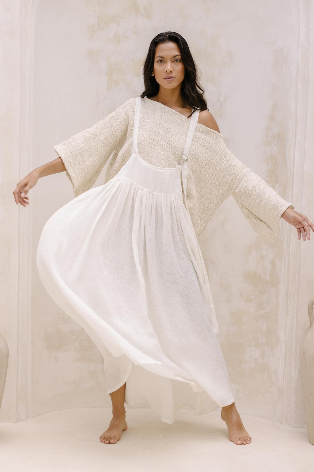 A woman with long dark hair poses barefoot against a neutral, textured background. She wears a loose-fitting, off-the-shoulder beige top and the May May Overall Skirt by Myrah Penaloza, a flowing white pinafore dress made to order from crinkle linen fabric. Her arms are gracefully extended, and the dress billows out around her as if she's in a magical fairy garden.