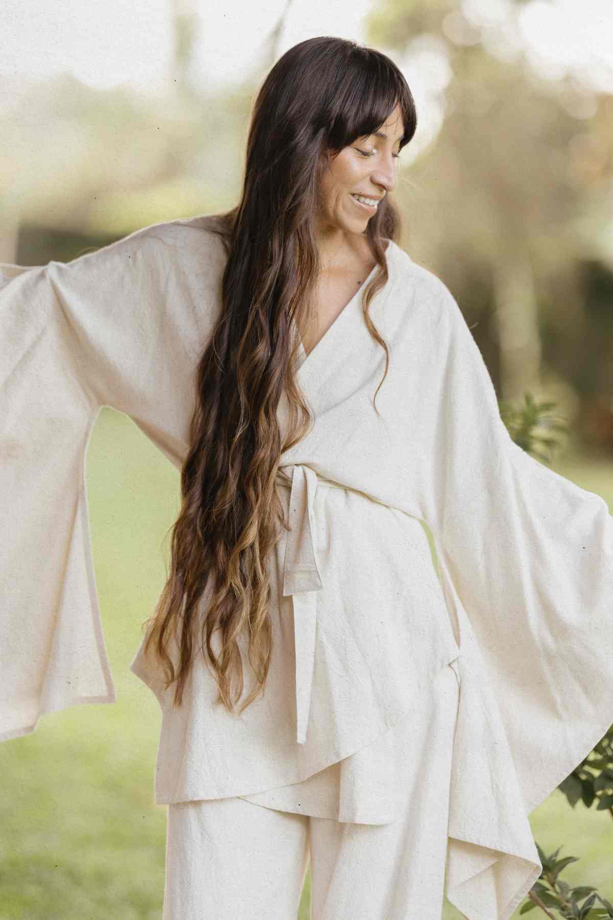 A woman with long, wavy brown hair smiles while looking down. She is wearing the light-colored Dewi Set (3 of a kind) - Pre-order by Myrah Penaloza, crafted from hypoallergenic linen cotton crepe with wide sleeves and a tied waist, standing outdoors with a green, blurred background.