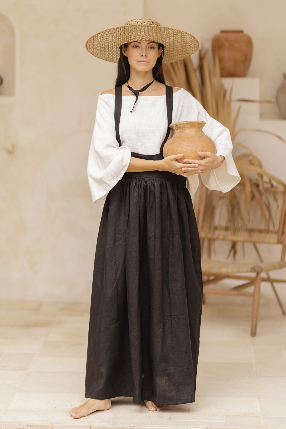 A person stands barefoot on a light-colored tile floor, wearing a wide-brimmed straw hat, a white off-the-shoulder blouse with loose sleeves, and the May May Overall Skirt (Made To Order) by Myrah Penaloza, crafted from 100% linen. They hold a clay pot in front of a neutral, earthy-toned background.