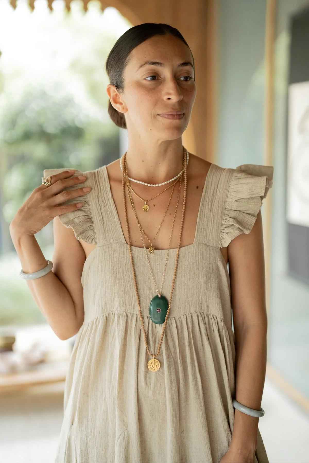 A woman with dark hair tied back in a sleek bun wears the Linen Anna Yemaya Gown from Myrah Penaloza. She accessorizes with multiple necklaces of varying lengths, including a green pendant, and bracelets. She stands in a light-filled room with a blurred background.