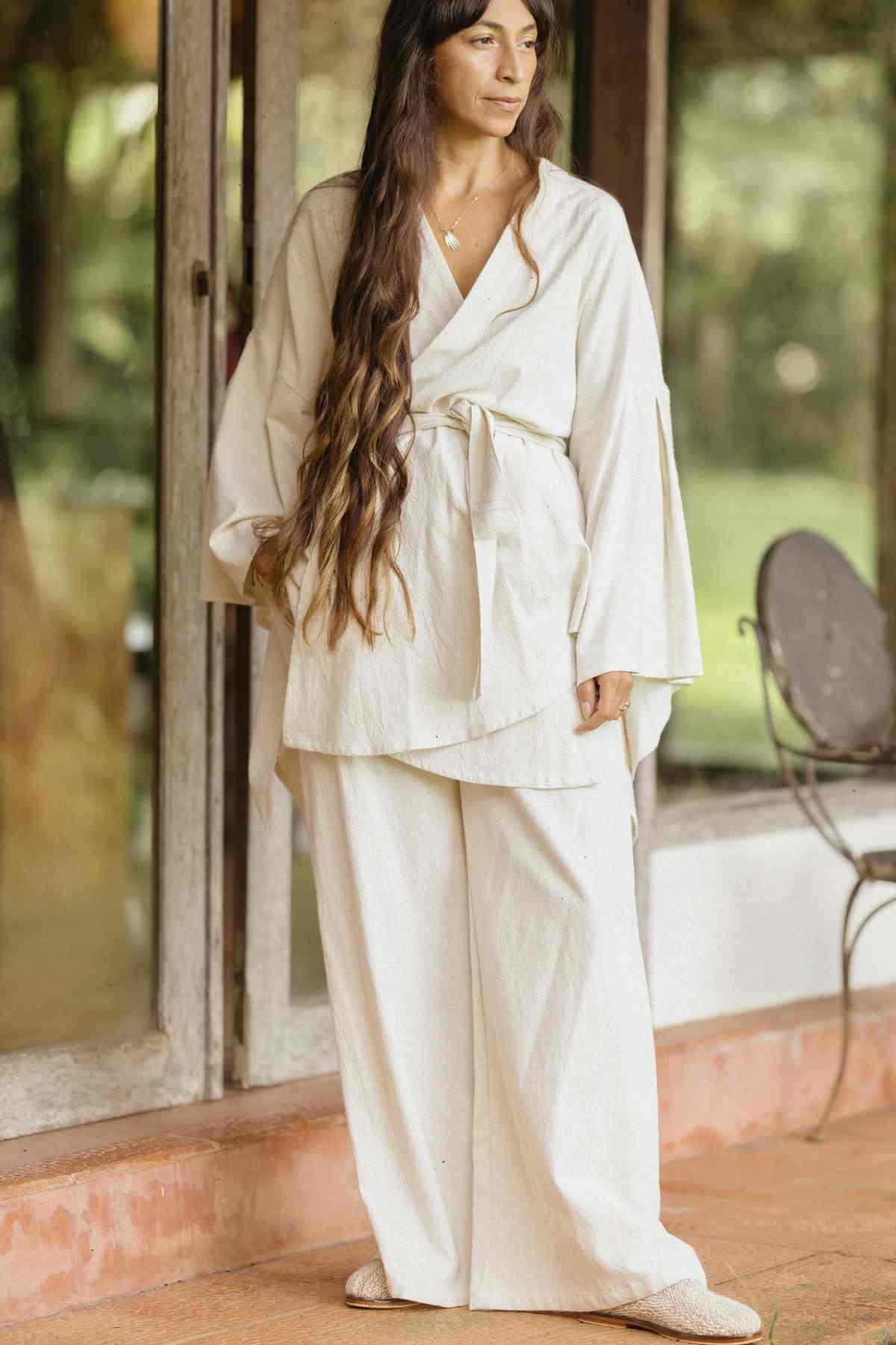 A woman with long, wavy hair stands on a porch wearing the Dewi Set (3 of a kind) by Myrah Penaloza—a light beige, belted kimono-style top paired with matching wide-leg pants crafted from hypoallergenic linen cotton crepe. The background showcases glass doors and a lush green outdoor setting, with a metal chair visible to the right.