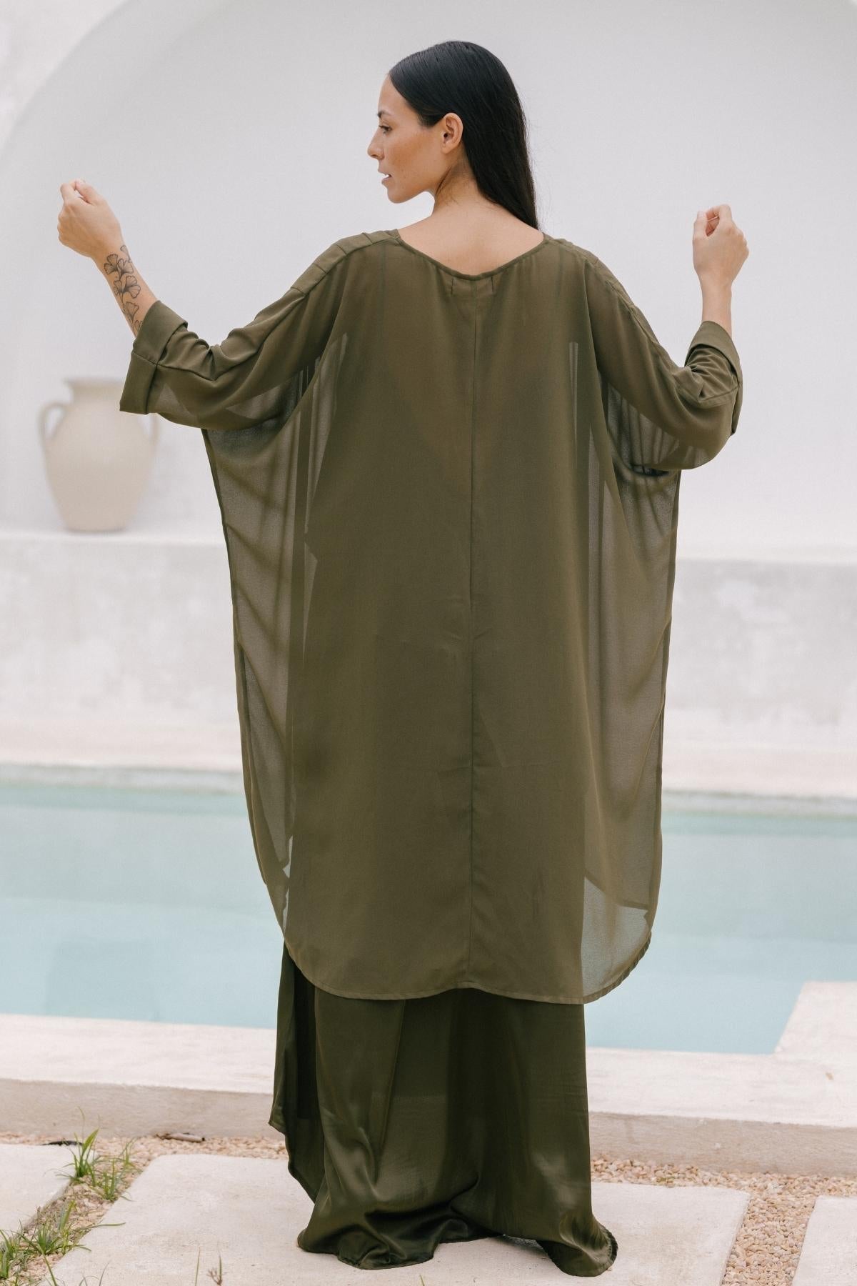 Standing by a pool, a person with long hair is draped in an olive green Cape made of 100% silk by Myrah Penaloza. With their back turned and arms slightly raised, the scene is complemented by a large vase against a white, curved wall—an embodiment of elegant style.