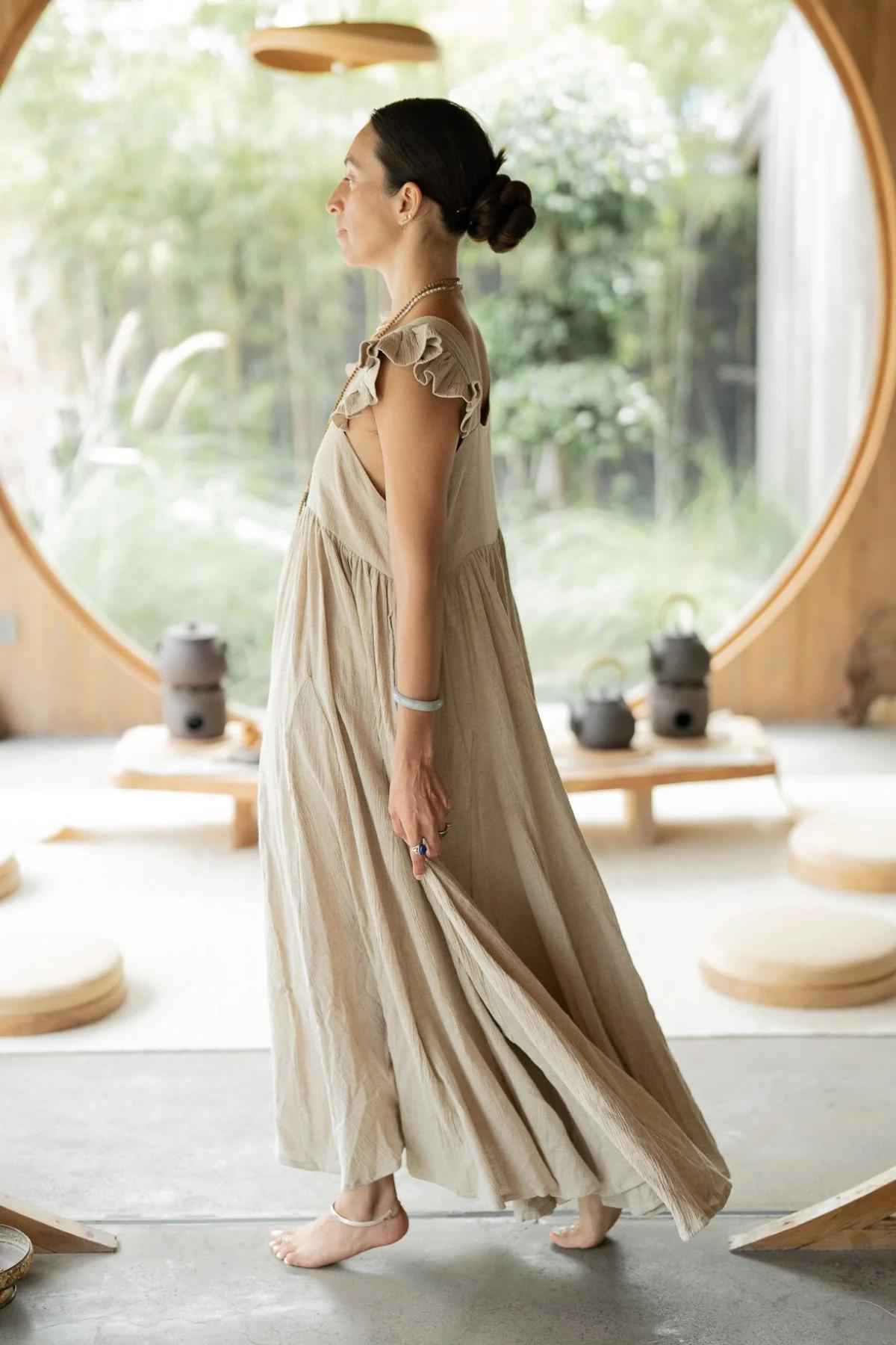 A woman in a long light beige Linen Anna Yemaya Gown by Myrah Penaloza walks barefoot in a minimalist room with large circular windows, allowing natural light to showcase the greenery outside. The room features low round tables and teapots, creating a serene and peaceful atmosphere.