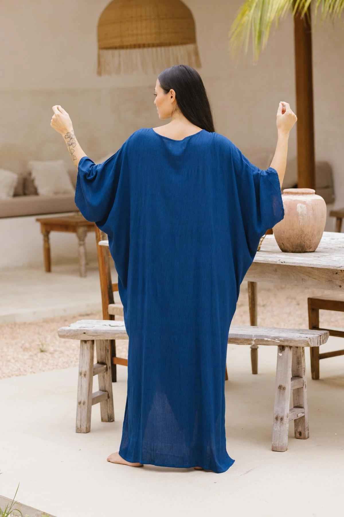 A person with long dark hair stands barefoot with their back to the camera, wearing a flowing, blue ankle-length Myrah Penaloza Limited Edition Virgo Kaftan. They hold their arms slightly away from their body. The setting includes a wooden table and benches, a large clay pot, and a hanging light fixture, exuding sophisticated fashion.