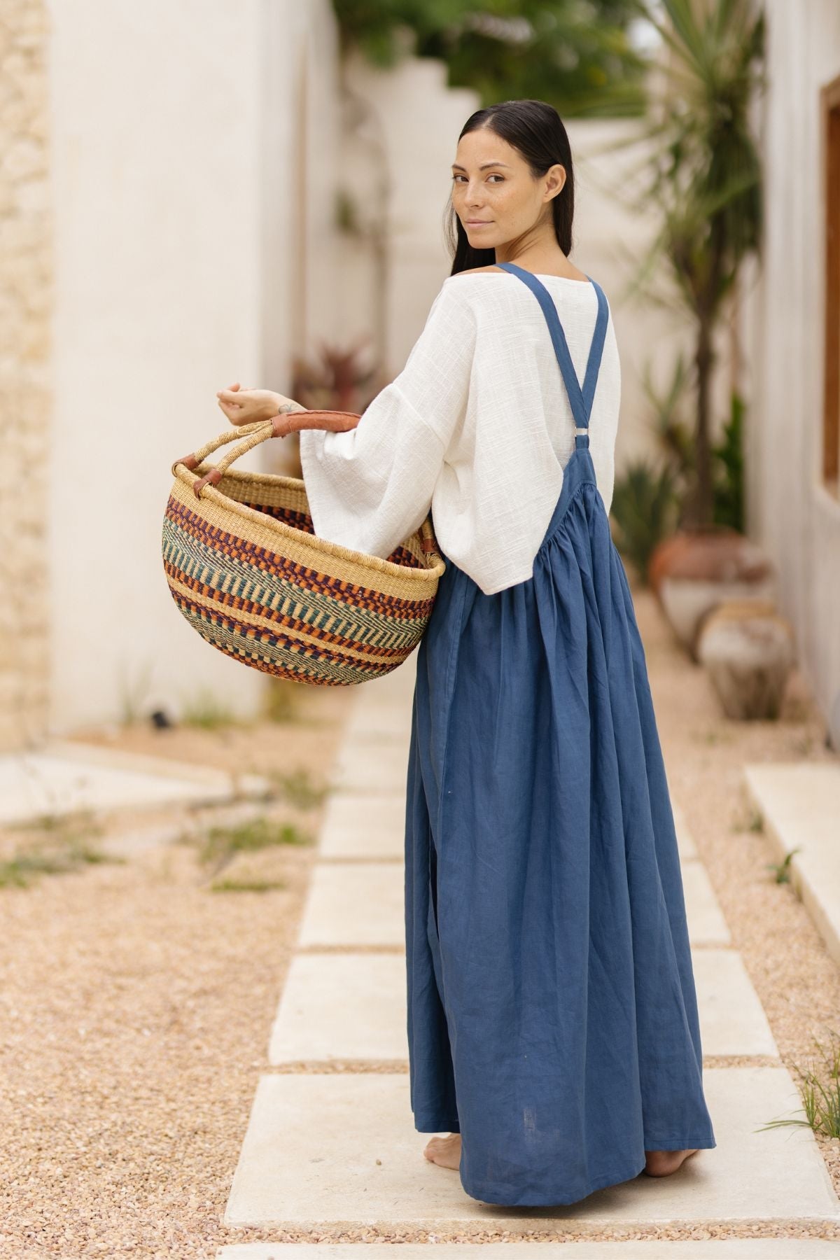 A woman with long dark hair stands on a gravel path, looking back over her shoulder. She wears a white blouse and the May May Overall Skirt (Made To Order) from Myrah Penaloza, made from crinkle linen fabric in a delightful blue hue. She is holding a large, colorful woven basket with both hands. The background includes potted plants and a white building, evoking the charm of a magical fairy garden.