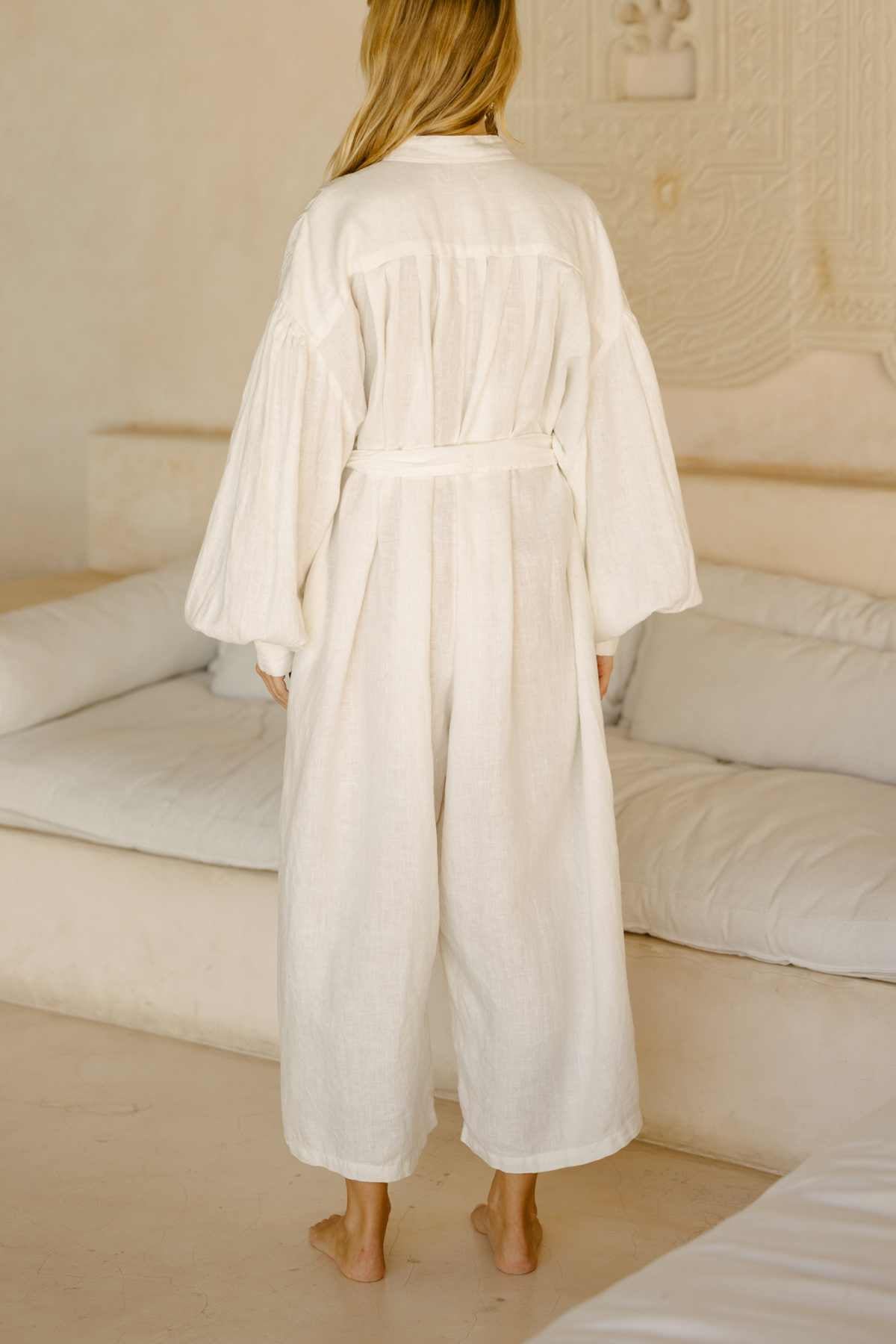 A person with long blond hair stands barefoot in a room featuring beige decor, wearing the Kundalini Gown Playsuit Edition by Myrah Penaloza. The 100% linen gown showcases billowy sleeves and a tie around the waist. The flowing white robe beautifully complements the serene atmosphere as they face away from the camera.