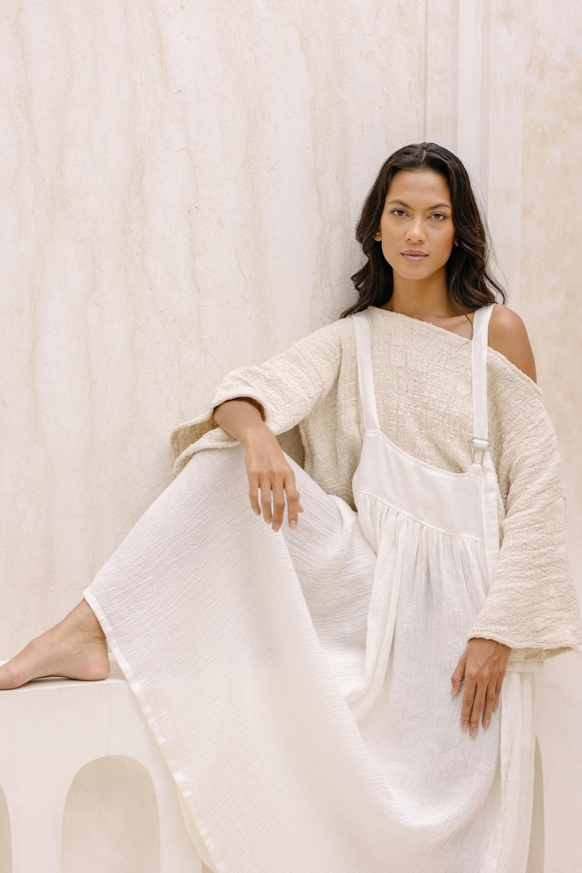 A person with long dark hair sits on a white bench against a light marble wall. They are wearing a beige off-the-shoulder sweater and the May May Overall Skirt by Myrah Penaloza, made of flowing, crinkle linen fabric. The person has a calm expression, their legs crossed with one foot resting on the bench, evoking the tranquility of a magical fairy garden.