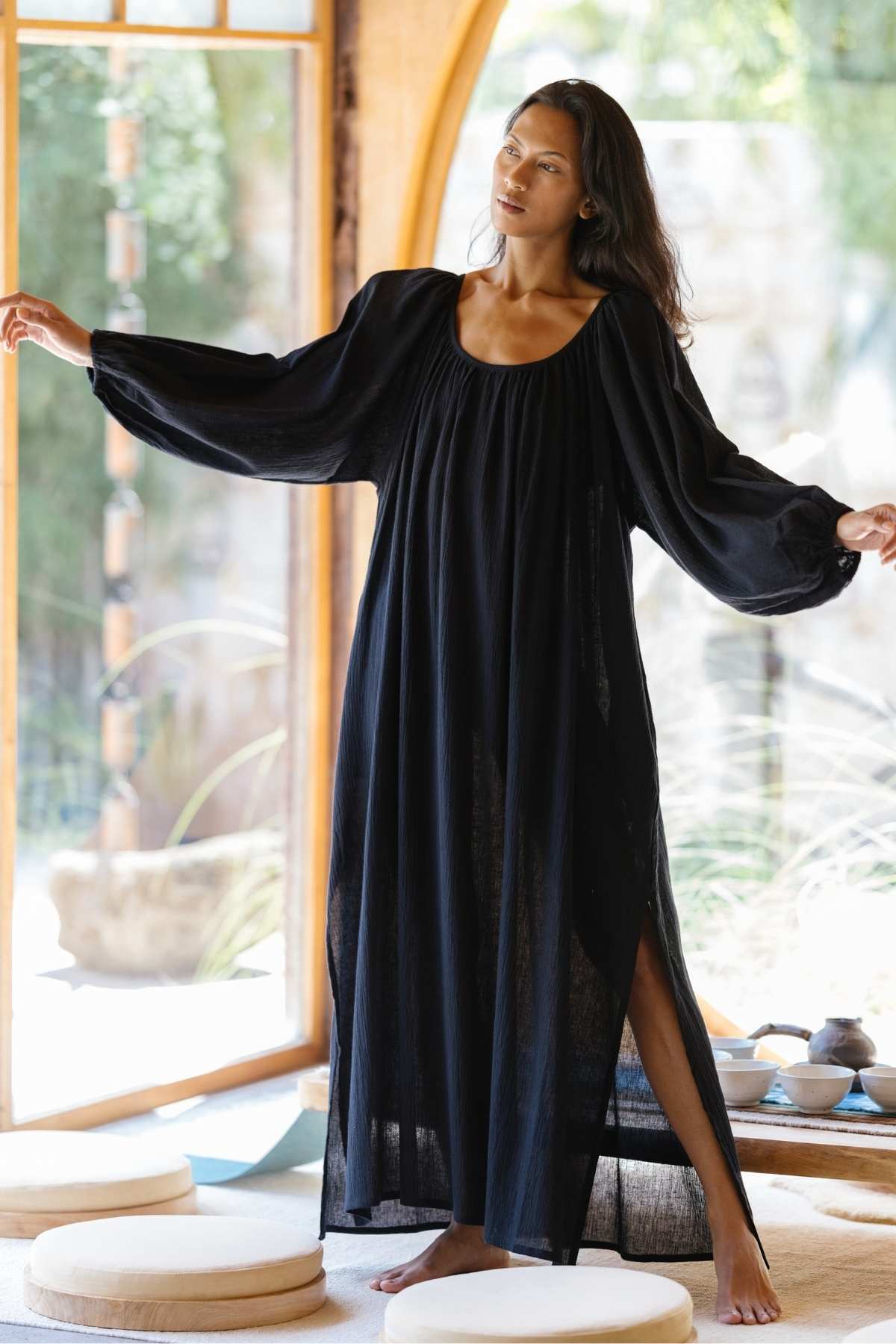 A woman with long hair stands barefoot in a room adorned with wooden decor and tall glass windows, wearing the flowing Alchemy Gown (Crinkle Linen Cotton) by Myrah Penaloza. Her long, billowy sleeves add to her graceful pose as she stretches her arms out, exuding a serene and modern mystic vibe.