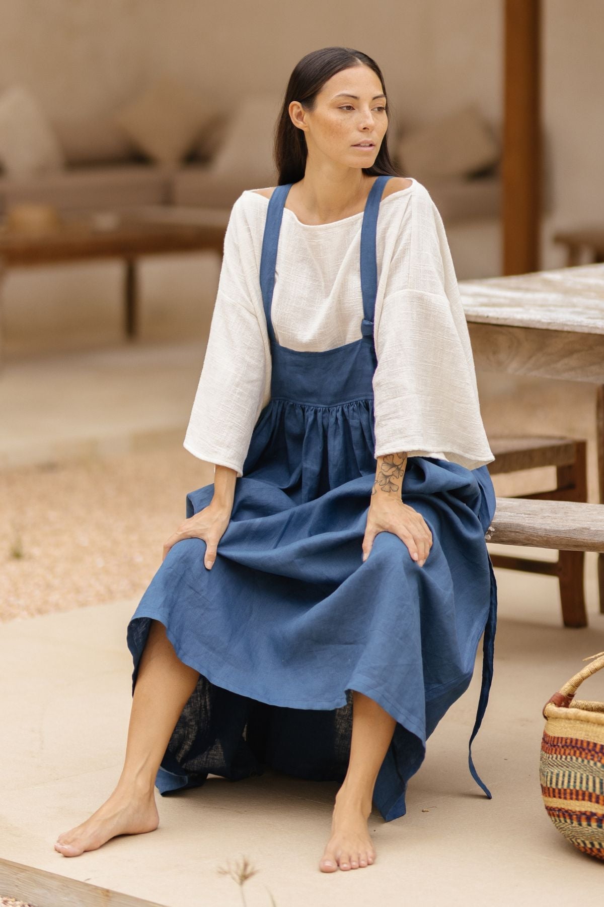 A woman with long dark hair sits on the edge of a wooden bench. She is wearing a loose white top with wide sleeves paired with the May May Overall Skirt, made of crinkle linen and custom-made by Myrah Penaloza. She is barefoot, and behind her, there is a woven basket and a rustic wooden table and bench.