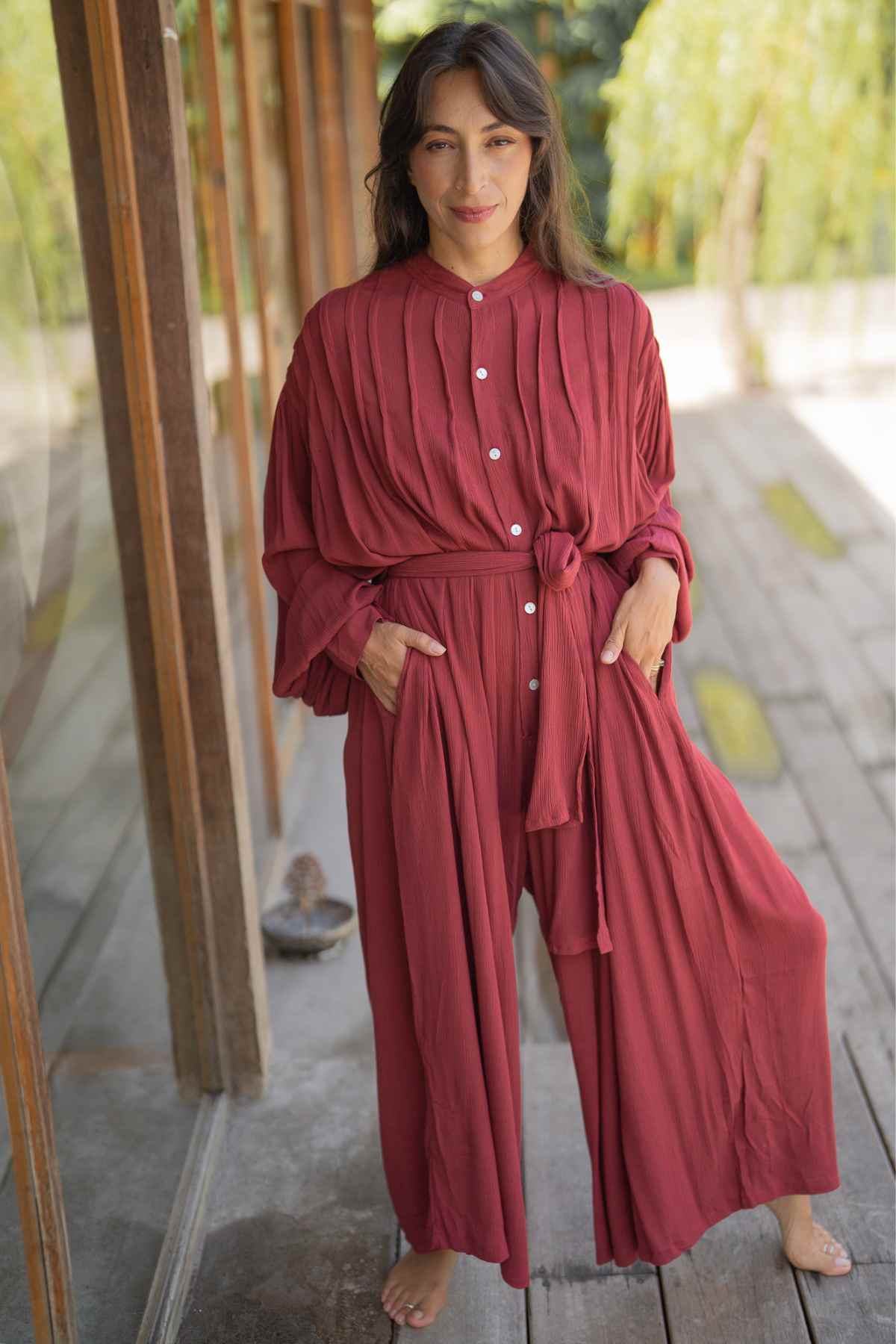 A person stands on a wooden walkway with greenery in the background. They are wearing a long, flowing burgundy dress similar to the Kundalini Gown Playsuit Edition by Myrah Penaloza, featuring buttons down the front. Their hands are in their pockets, and they are barefoot.