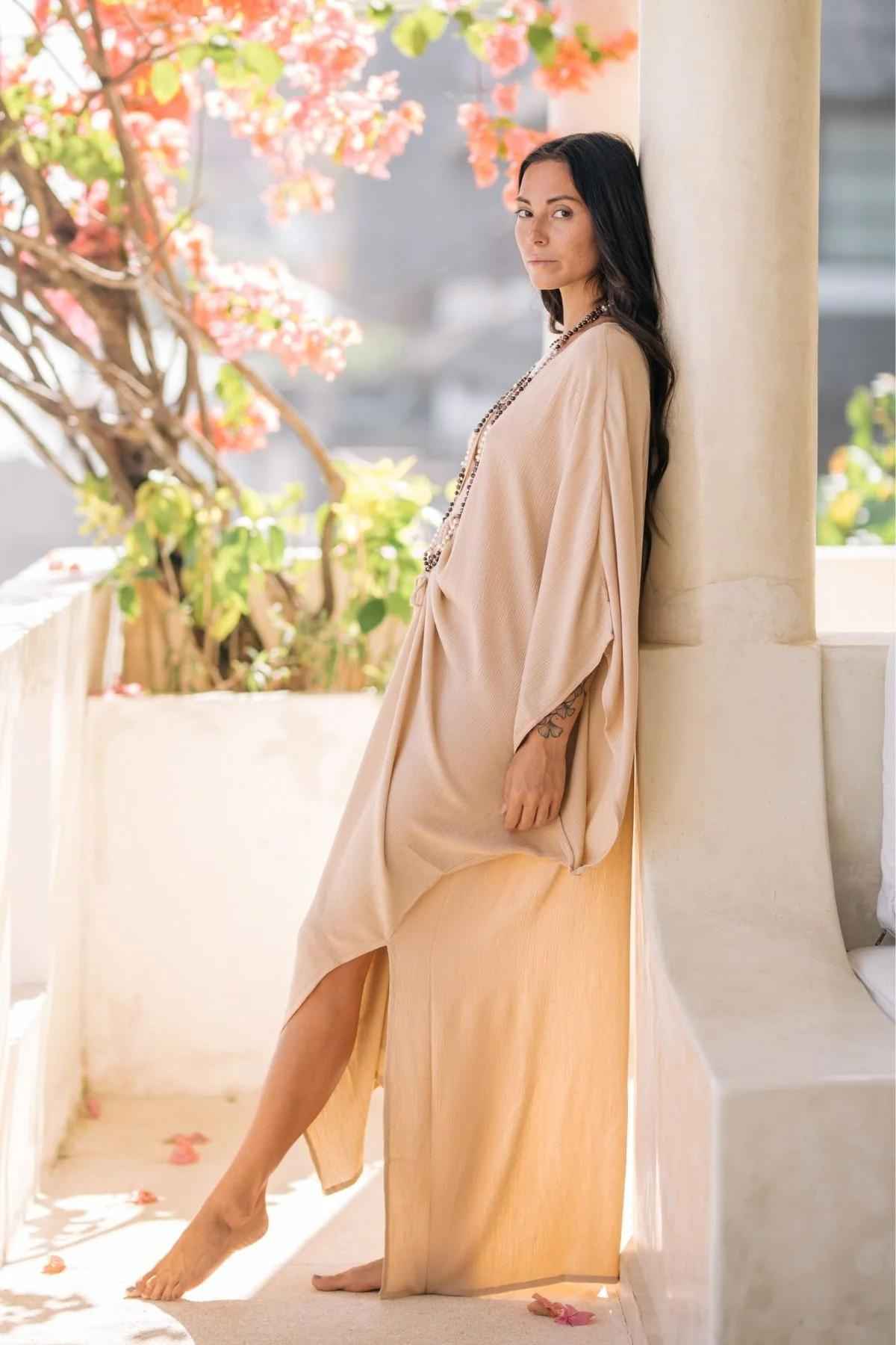 A woman in a beige long dress stands barefoot against a column on a bright, sunny day. She is outdoors on a white terrace adorned with pink flowers and green plants. Wearing the Limited Edition Virgo Kaftan from Myrah Penaloza, the sunlight casts soft shadows, creating a serene and peaceful atmosphere.