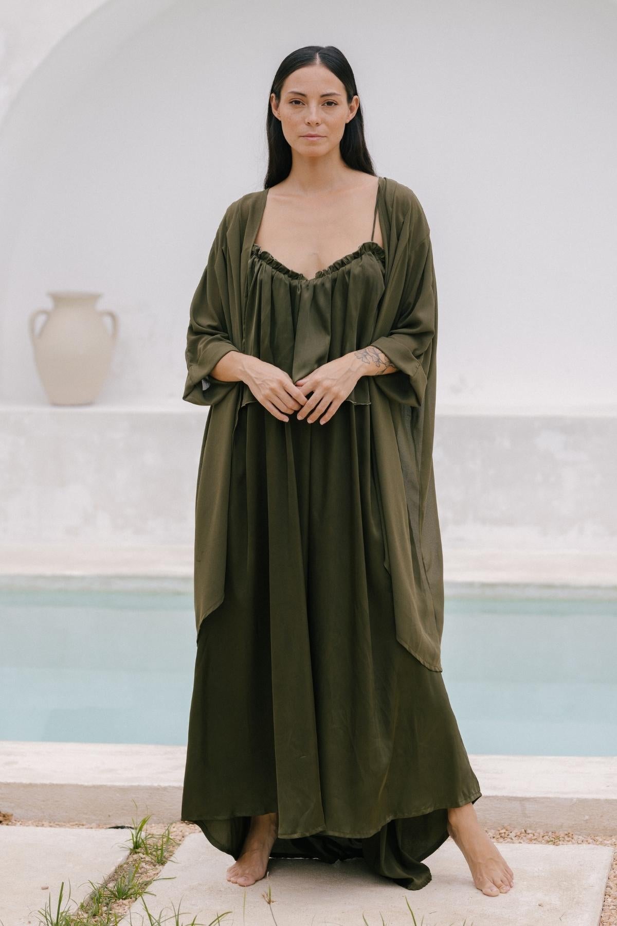 A woman stands by a pool wearing an exquisite olive-green cape crafted from 100% silk by Myrah Penaloza, her hands gently clasped in front. An elegant urn sits in the background against a white wall, enhancing the serene setting.