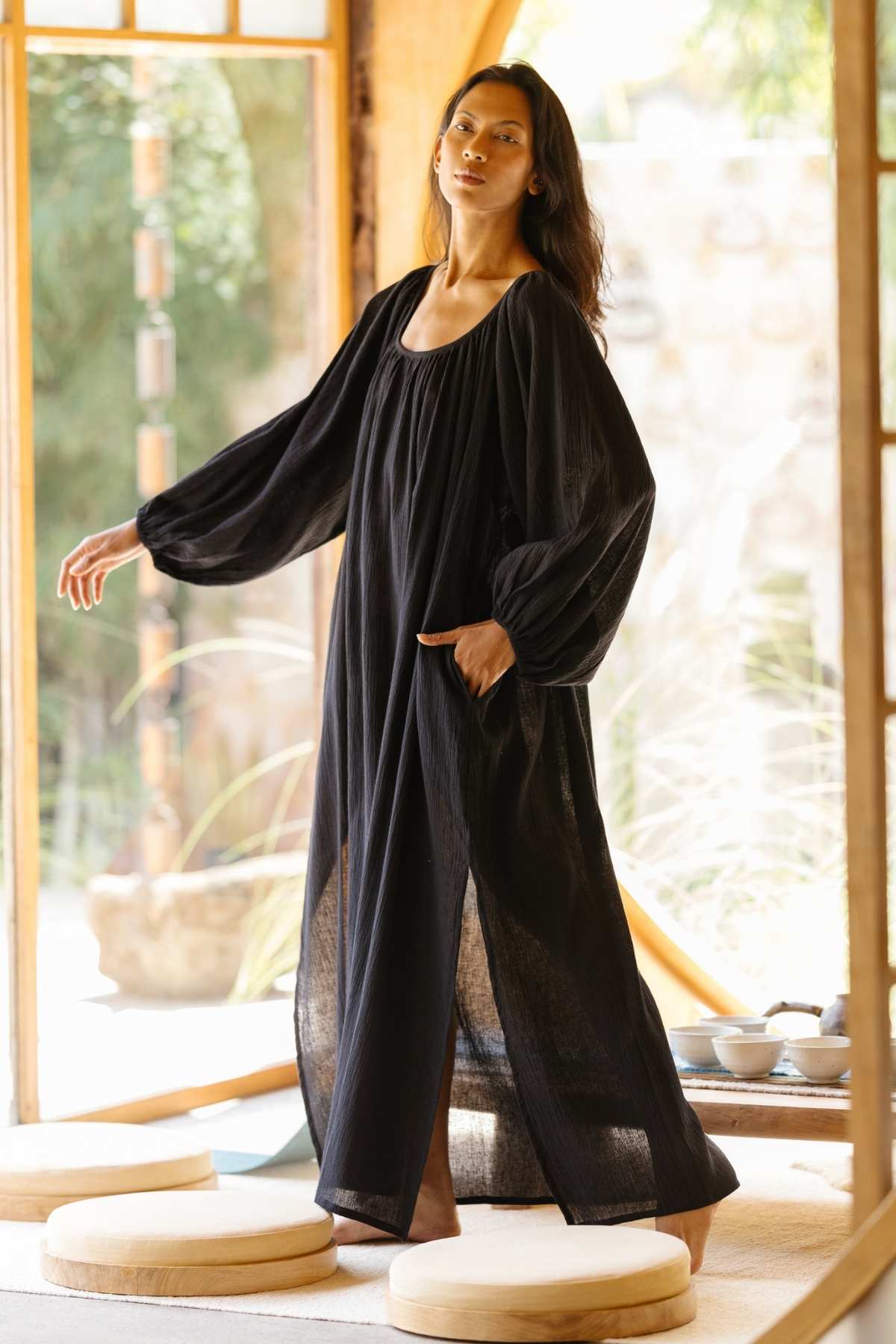 A person stands indoors in a spacious, well-lit room. They are wearing a long, flowing Alchemy Gown (Crinkle Linen Cotton) by Myrah Penaloza with voluminous sleeves and have long hair. The background includes large windows, wooden doors, round cushions on the floor, and a blurred outdoor scene, adding to the modern mystic vibe.