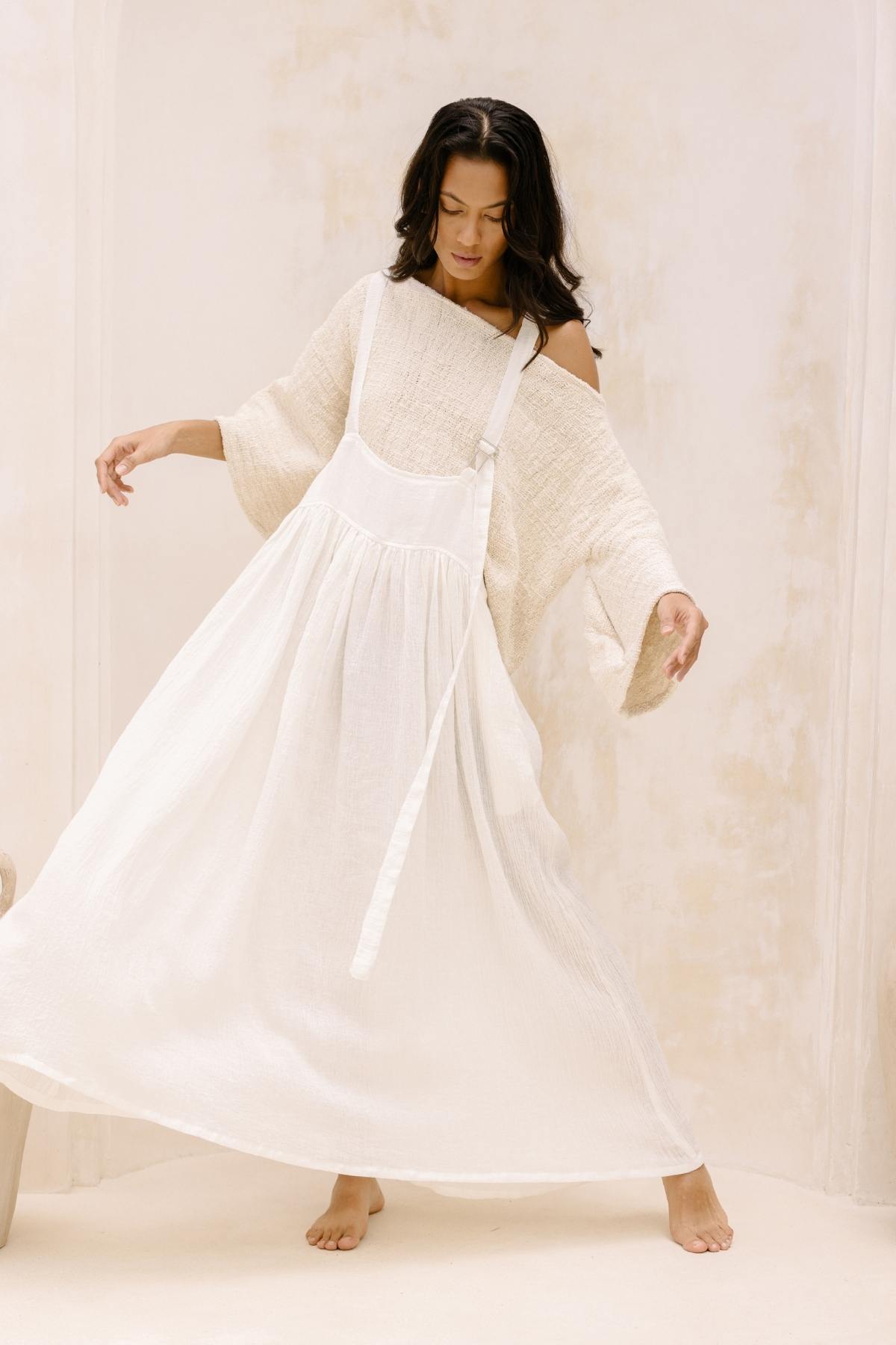 A woman wearing a loose, off-the-shoulder white top and the Myrah Penaloza May May Overall Skirt (Made To Order) poses in a softly lit, neutral-toned room. Her dark hair is down, and she appears to be in mid-movement with her arms slightly extended, creating a graceful and flowing effect.