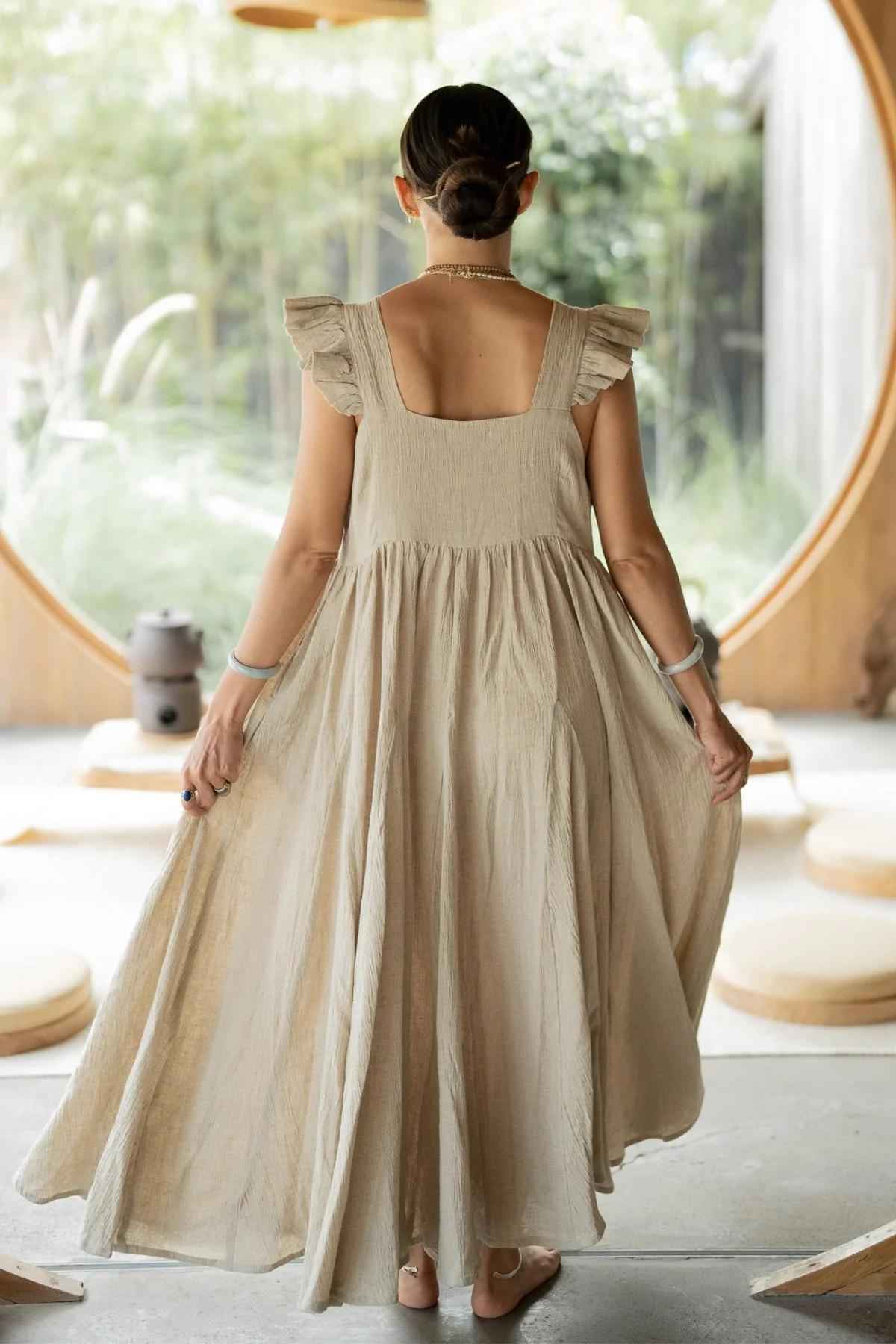 A woman with her hair in a bun stands with her back to the camera, wearing a long, flowing Linen Anna Yemaya Gown from Myrah Penaloza, featuring ruffled sleeves made of crinkle cotton. She is in a minimalist indoor setting with round wooden structures and green scenery visible through a large circular window.