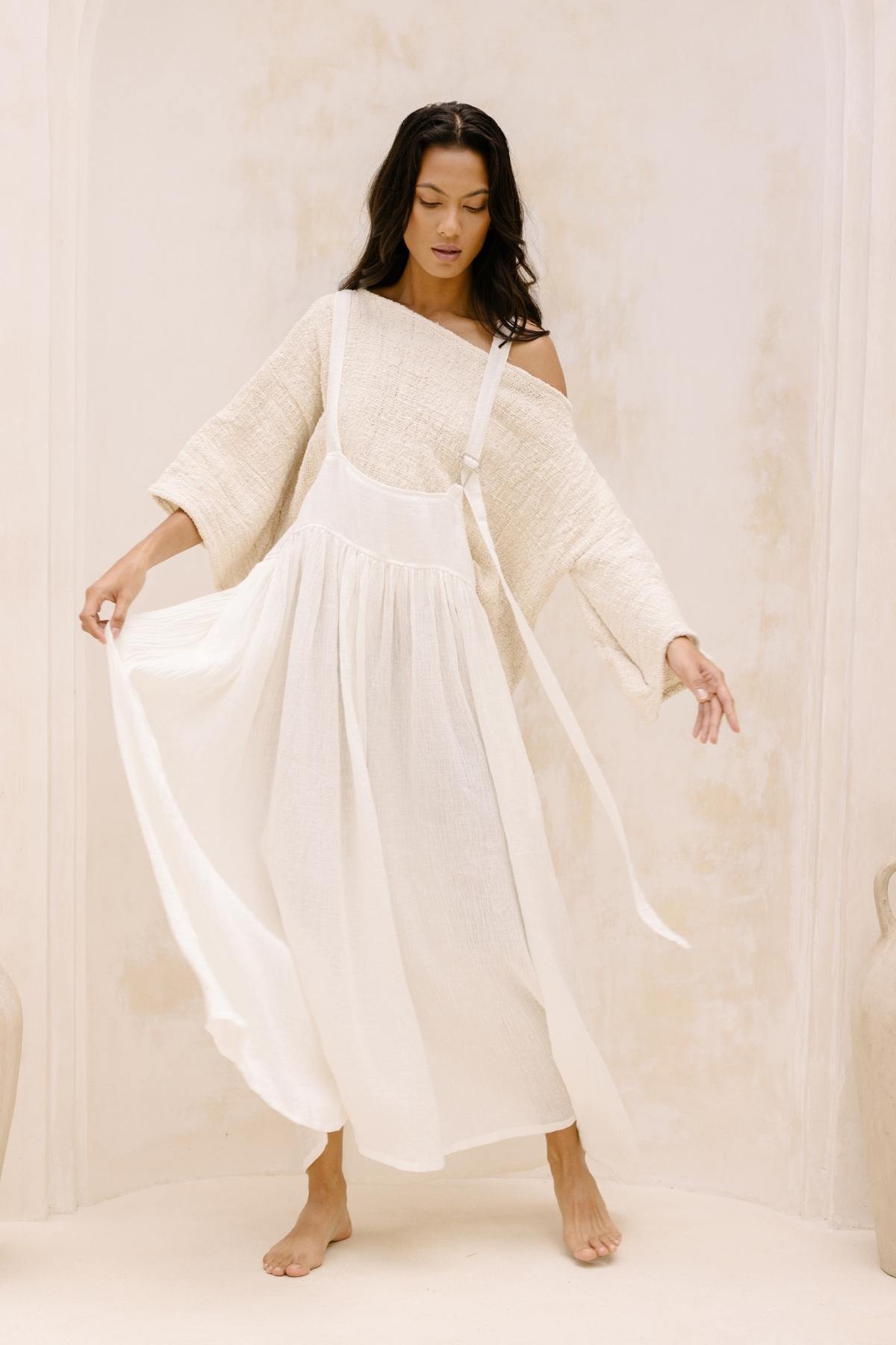 A woman stands barefoot against an off-white background, wearing an off-the-shoulder, long-sleeved, beige top and the May May Overall Skirt by Myrah Penaloza. Her eyes are downcast as she delicately holds the sides of her flowing white skirt, creating a sense of gentle motion.