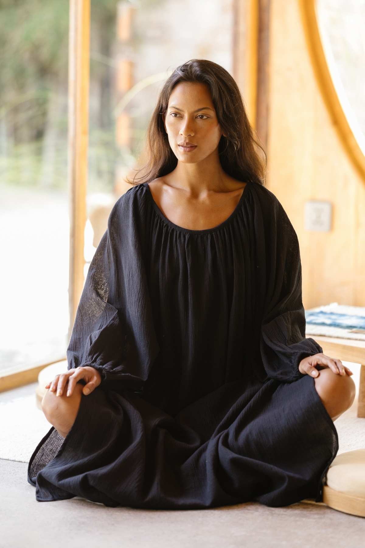 A woman with long, dark hair sits cross-legged on a cushion near a large window. She wears an elegant Alchemy Gown (Crinkle Linen Cotton) by Myrah Penaloza, its long sleeves adding to her graceful appearance. The background reveals a bright, natural outdoor setting with trees. She appears calm and centered, possibly meditating, embodying the essence of a modern mystic.