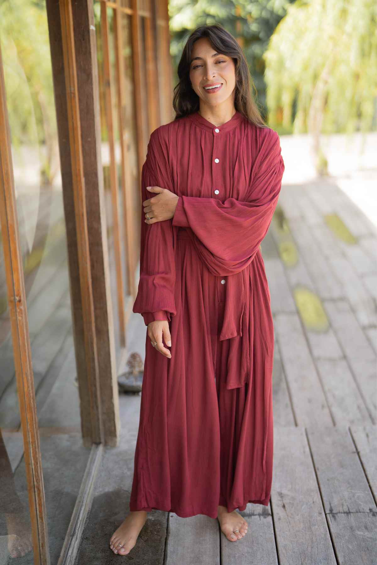 A woman stands barefoot on a wooden deck beside a tall window, wearing the long, flowing Kundalini Gown Playsuit Edition from Myrah Penaloza. Her long, wavy hair cascades gently as she smiles softly, looking slightly to the side. The background is blurred greenery.