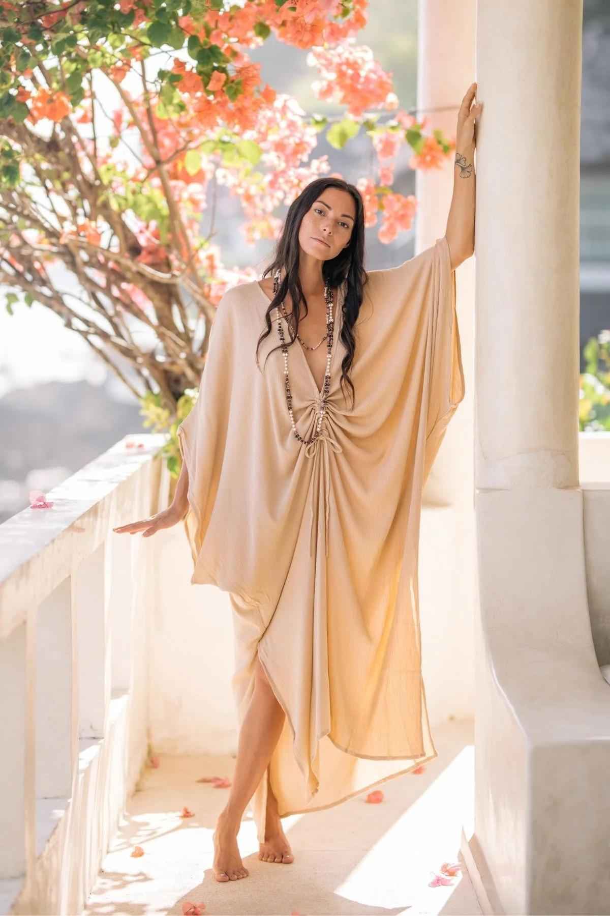 A woman stands barefoot on a sunlit outdoor walkway, leaning against a column. She is wearing a flowing, light beige Limited Edition Virgo Kaftan from Myrah Penaloza, accessorized with long necklaces. Behind her are branches with bright pink flowers. The scene is serene and bright.