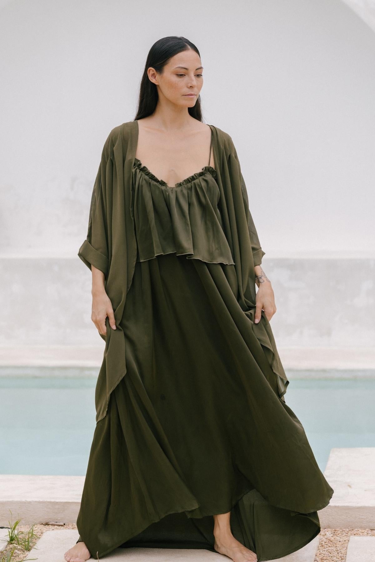 A person in a flowing olive green dress with an elegant style stands barefoot by a pool, their long dark hair cascading down. A luxurious 100% silk cape by Myrah Penaloza enhances their serene and relaxed vibe. The background features a white wall and shallow water in the pool.