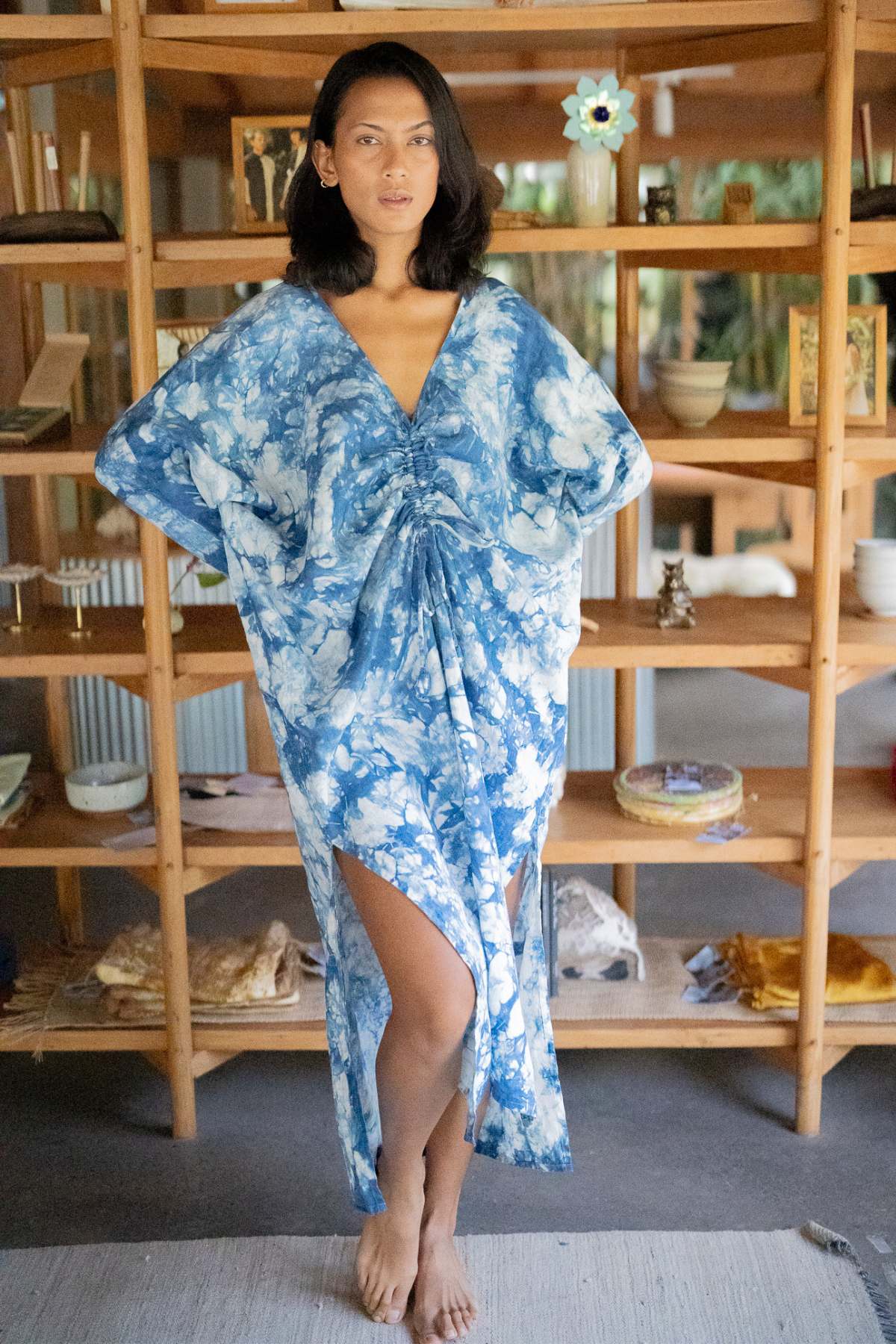 A person stands barefoot in front of a wooden shelf, dazzling in the exclusive Indigo Virgo Rising Kaftan by Myrah Penaloza. This blue and white tie-dye masterpiece features a ruched front with a high slit, while the background is adorned with decorative items and lush plants.