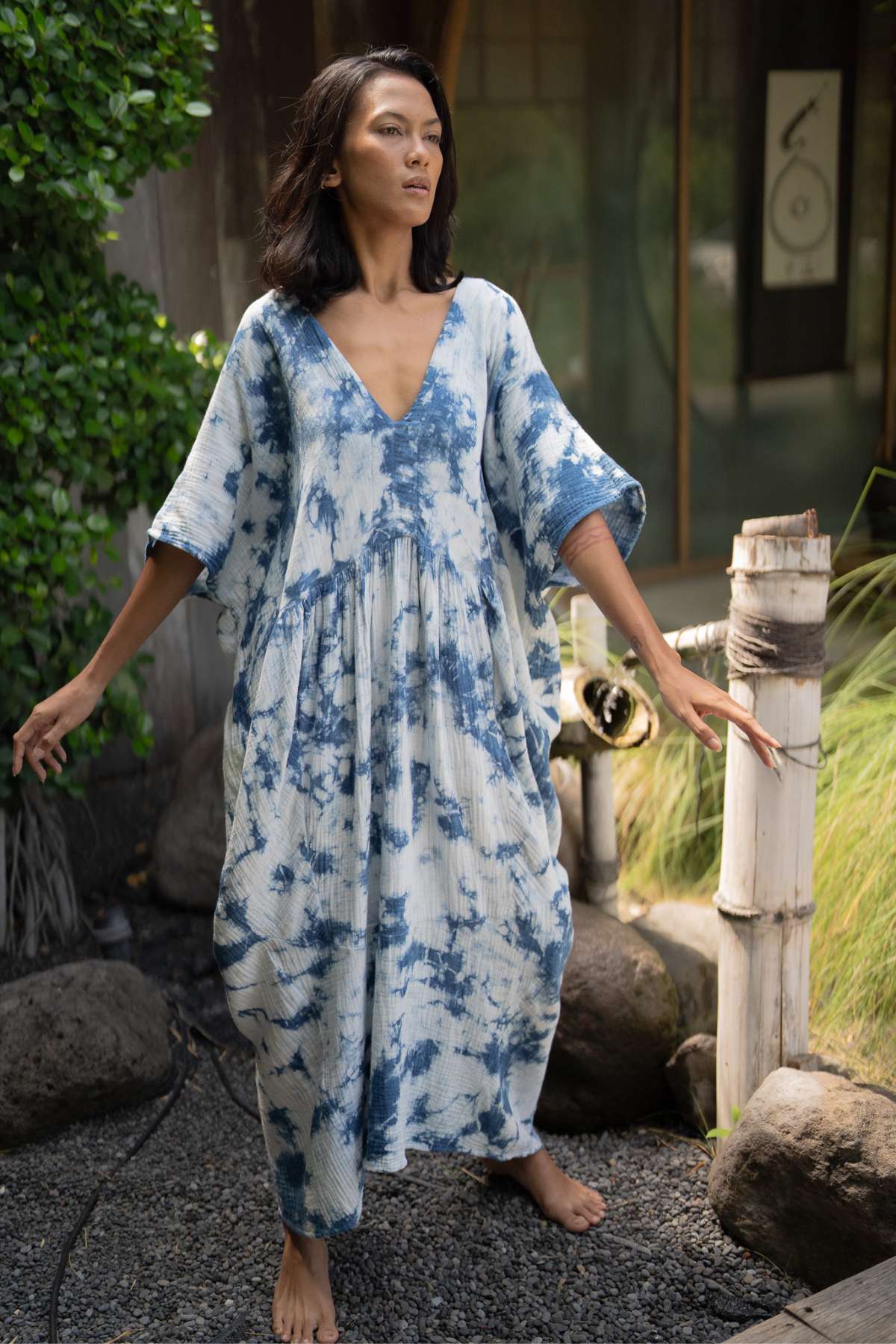 A person stands outdoors on a gravel path, barefoot and surrounded by greenery and wooden posts, exuding an effortless style in the Indigo Venus Gown by Myrah Penaloza. The flowing cotton gauze gown features a deep V-neck and blue and white tie-dye design, perfectly capturing the serene essence of nature, reminiscent of Venus herself.