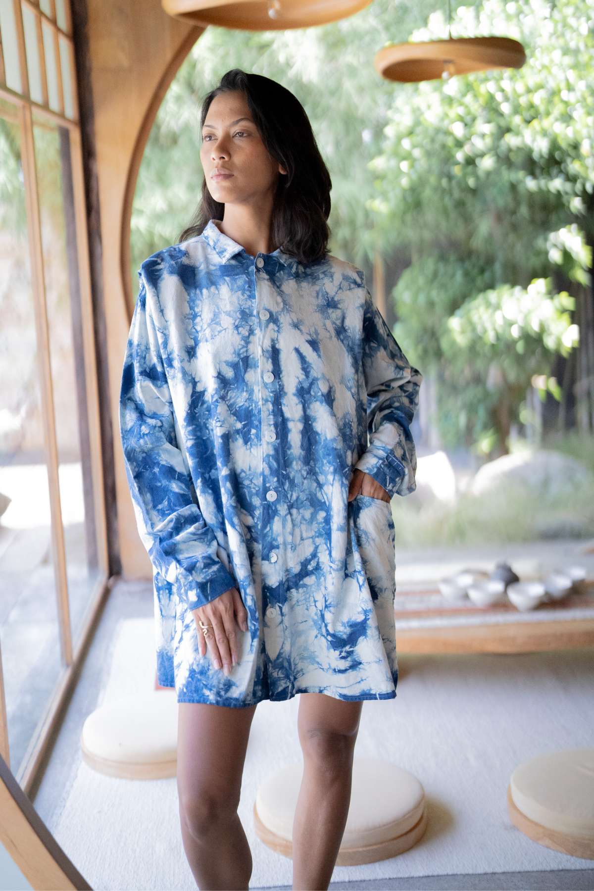 A person with shoulder-length dark hair stands indoors near a large window, wearing the Tender Jacket - Hand Tye Dyed Indigo by Myrah Penaloza. They gaze thoughtfully into the distance, surrounded by wooden accents and a view of greenery outside.