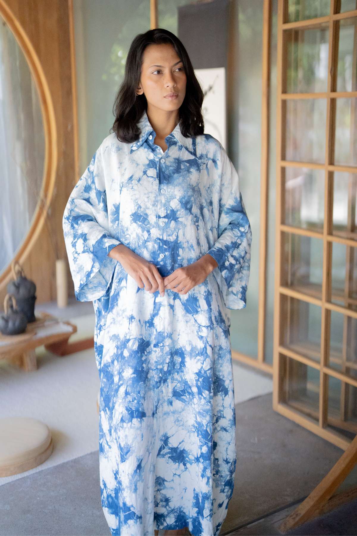 A woman with long dark hair stands in a room wearing The Boyfriend Dress - Hand Tie Dyed Indigo by Myrah Penaloza. She is positioned near a wooden sliding door adorned with circular patterns, creating a calm and stylish atmosphere.