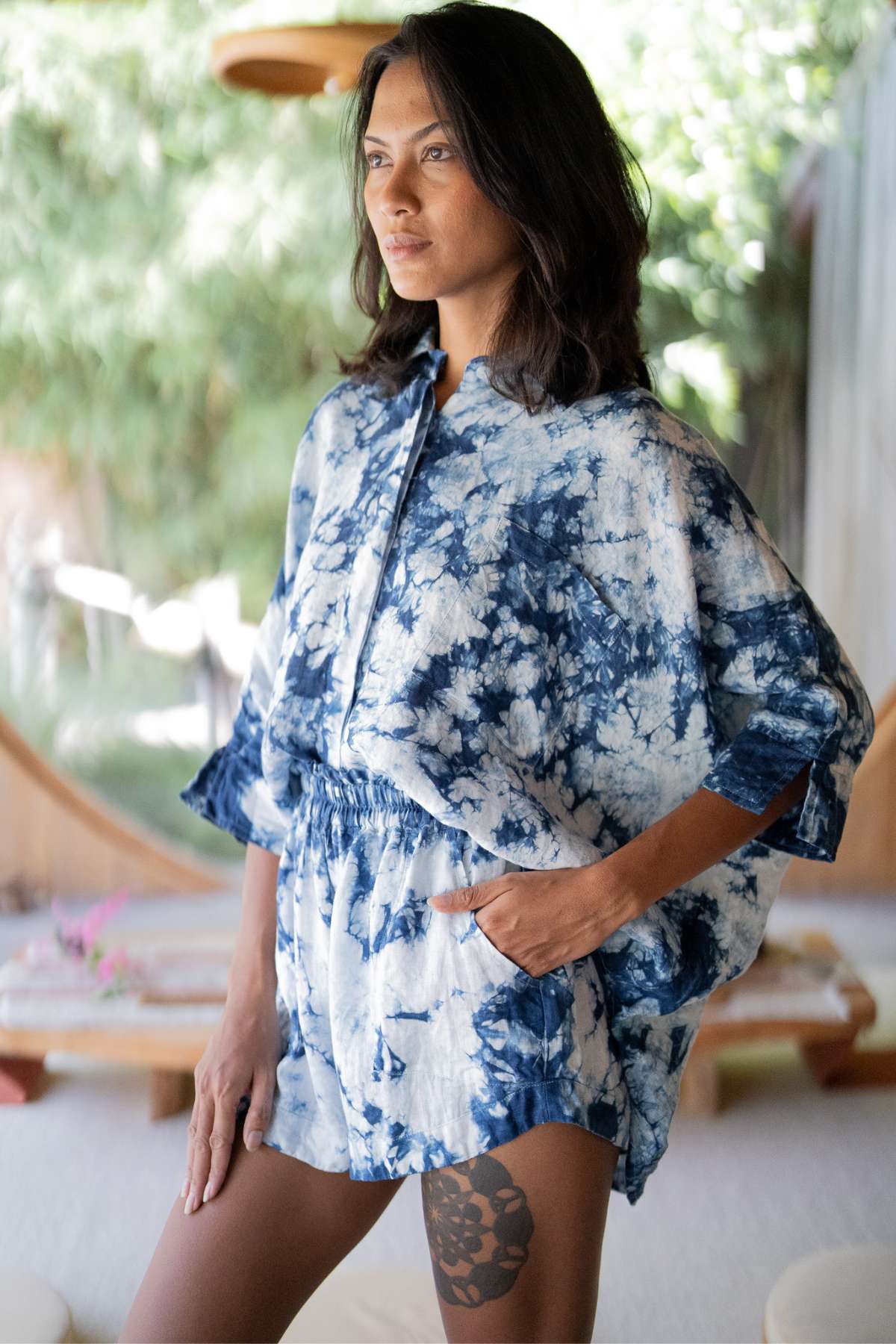 A person stands indoors wearing the Indigo Suka Set by Myrah Penaloza, a blue and white tie-dye outfit made from ethically sourced 100% linen, consisting of a loose top and shorts. They have long dark hair and a tattoo on their thigh. The background is softly blurred with natural light shining through greenery.