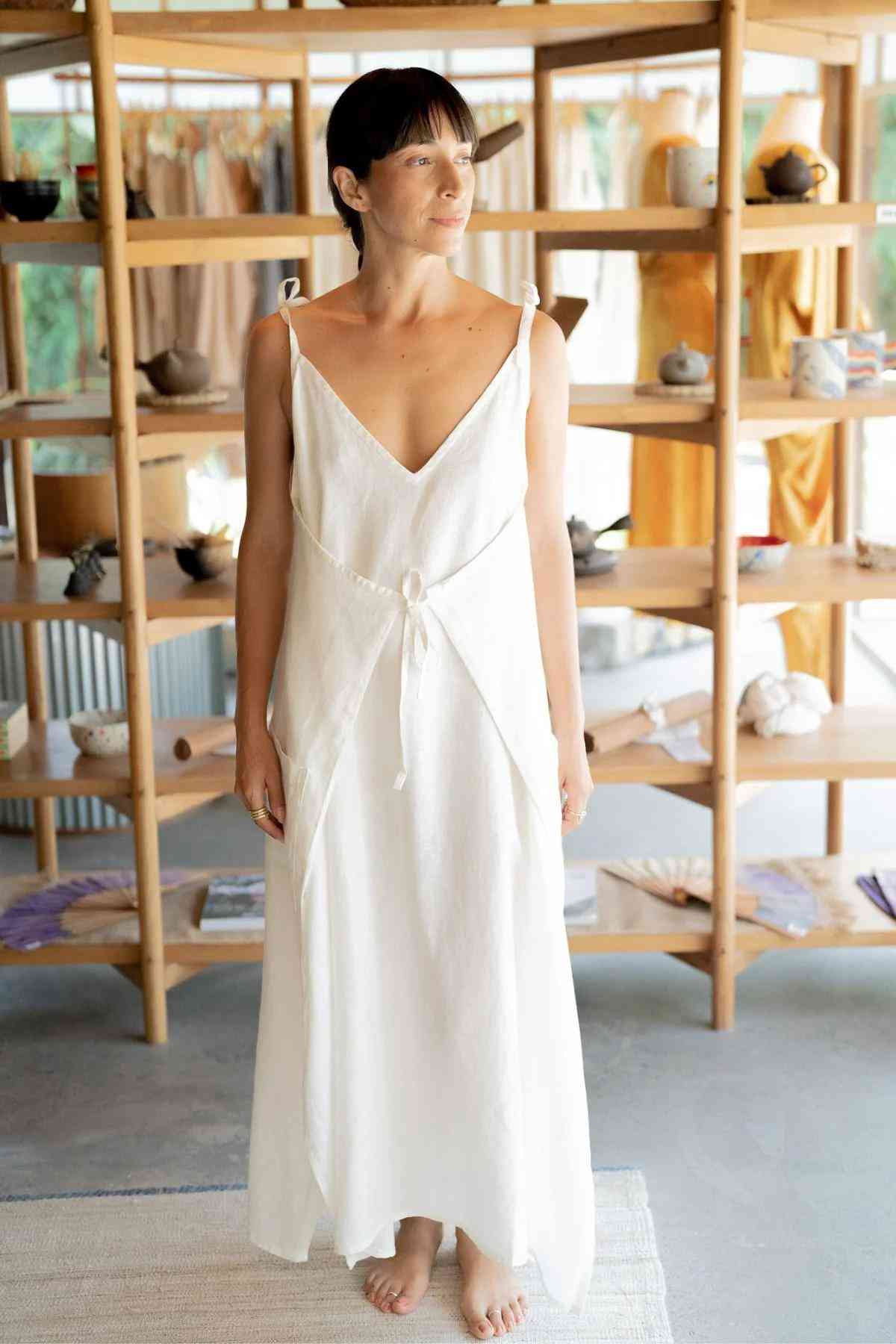 A woman stands barefoot on a smooth light grey floor, adorned in the Radiant Gown - Made To Order by Myrah Penaloza from the healing linen collection. The long, sleeveless white dress with thin straps tied at her shoulders exudes elegance. Behind her, wooden shelves display various pottery items and colorful clothing in the background.
