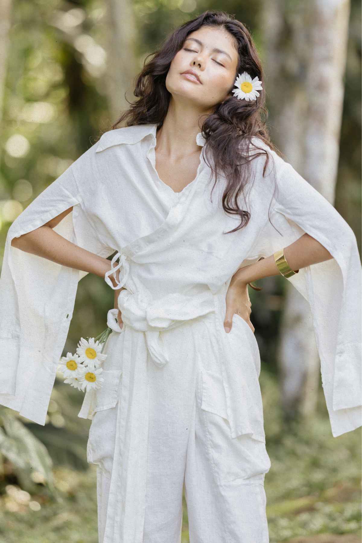 A woman with long dark hair stands outdoors, donning the versatile and lightweight Sutra Linen Set (Made To Order) by Myrah Penaloza. Daisies are tucked into her hair and pocket, complementing her serene expression and closed eyes. The stylish and sustainable background fades into blurred greenery, enhancing the tranquil scene.