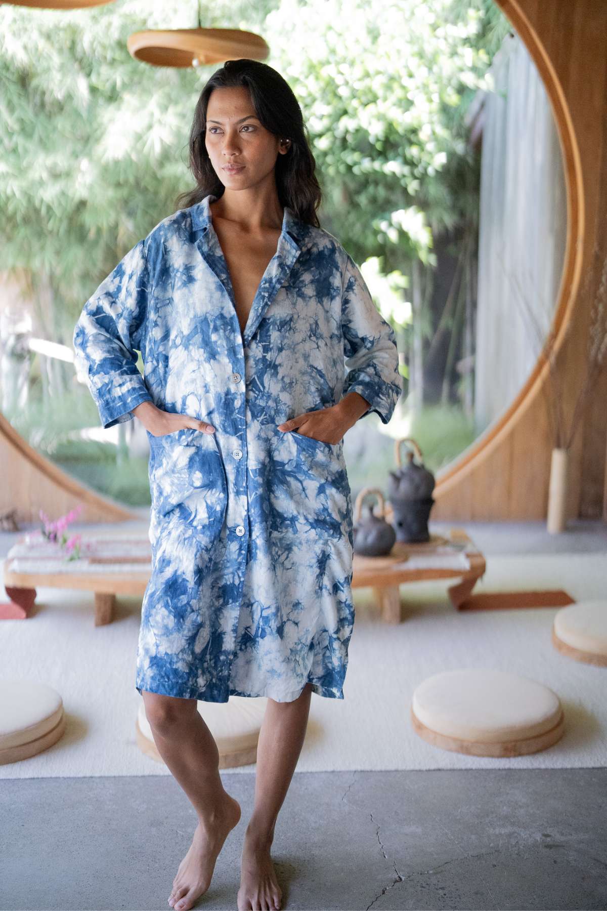 A woman stands barefoot in a room with a large circular window, wearing the Rubi Linen Jacket in Indigo from Myrah Penaloza, with her hands casually resting in the pockets. The scene is set against a backdrop of cushions, teapots on a table, and lush green foliage visible through the window.