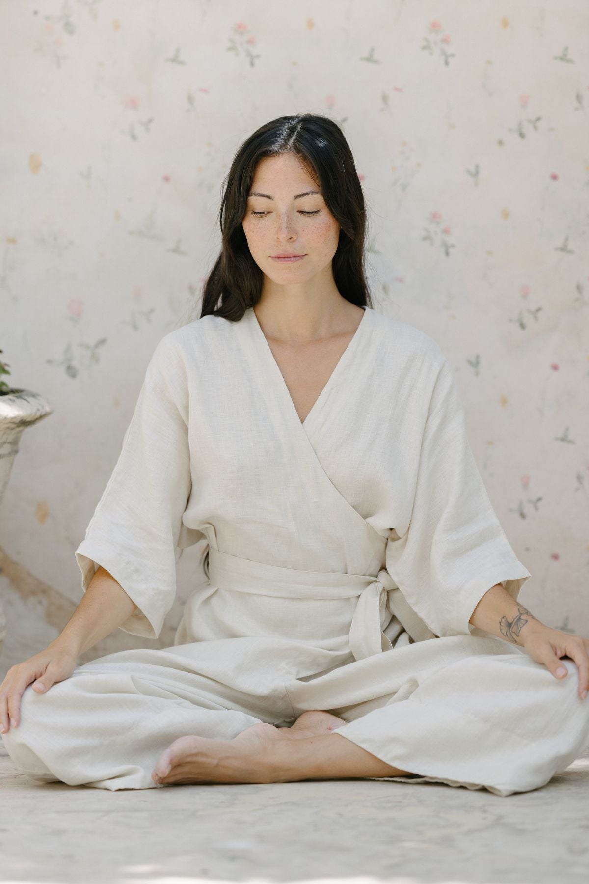 A person with long dark hair is sitting cross-legged on the floor, wearing the Rey Linen Playsuit by Myrah Penaloza, which features a light-colored kimono-style design with long sleeves and an adjustable front wrap. Their eyes are closed, and their hands rest on their knees. The background is softly patterned with delicate floral designs.