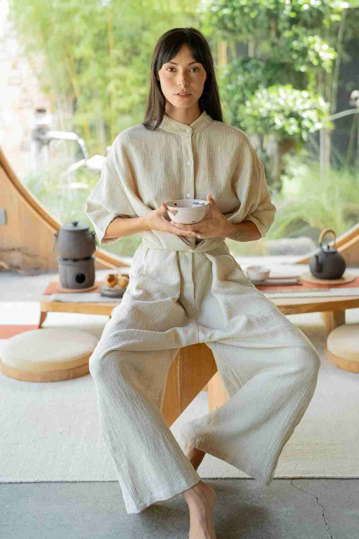 A person with long hair, dressed in Myrah Penaloza's relaxed, light-colored Pine Playsuit, sits cross-legged on a wooden bench holding a bowl. Behind them, there is a round window revealing greenery outside. The setting includes a low table with tea accessories—a scene of serene simplicity.