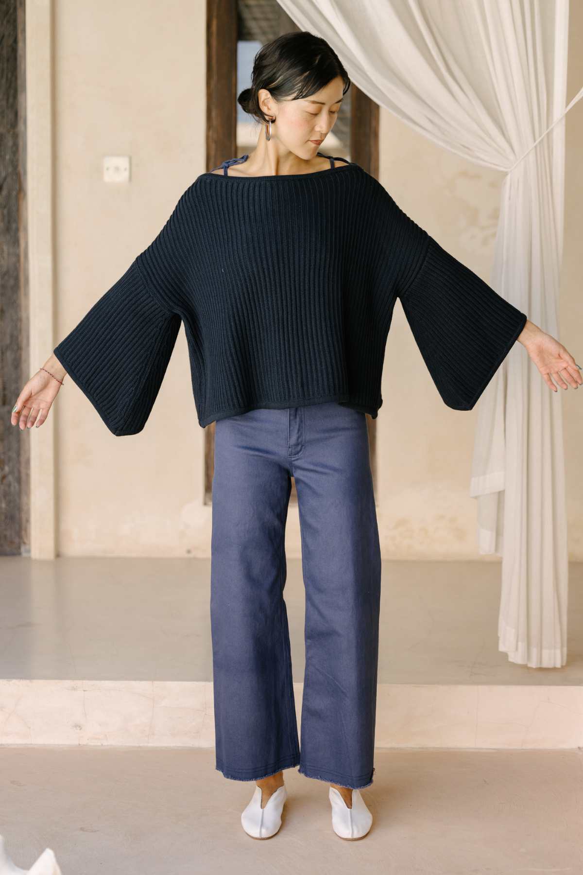 A person with short dark hair stands indoors with arms outstretched, showcasing the oversized black Spirit Top Sweater Knit made by Myrah Penaloza, crafted from luxurious cotton fabric. They pair it with wide-leg blue jeans against a backdrop of a beige wall featuring a window draped in sheer white curtains.