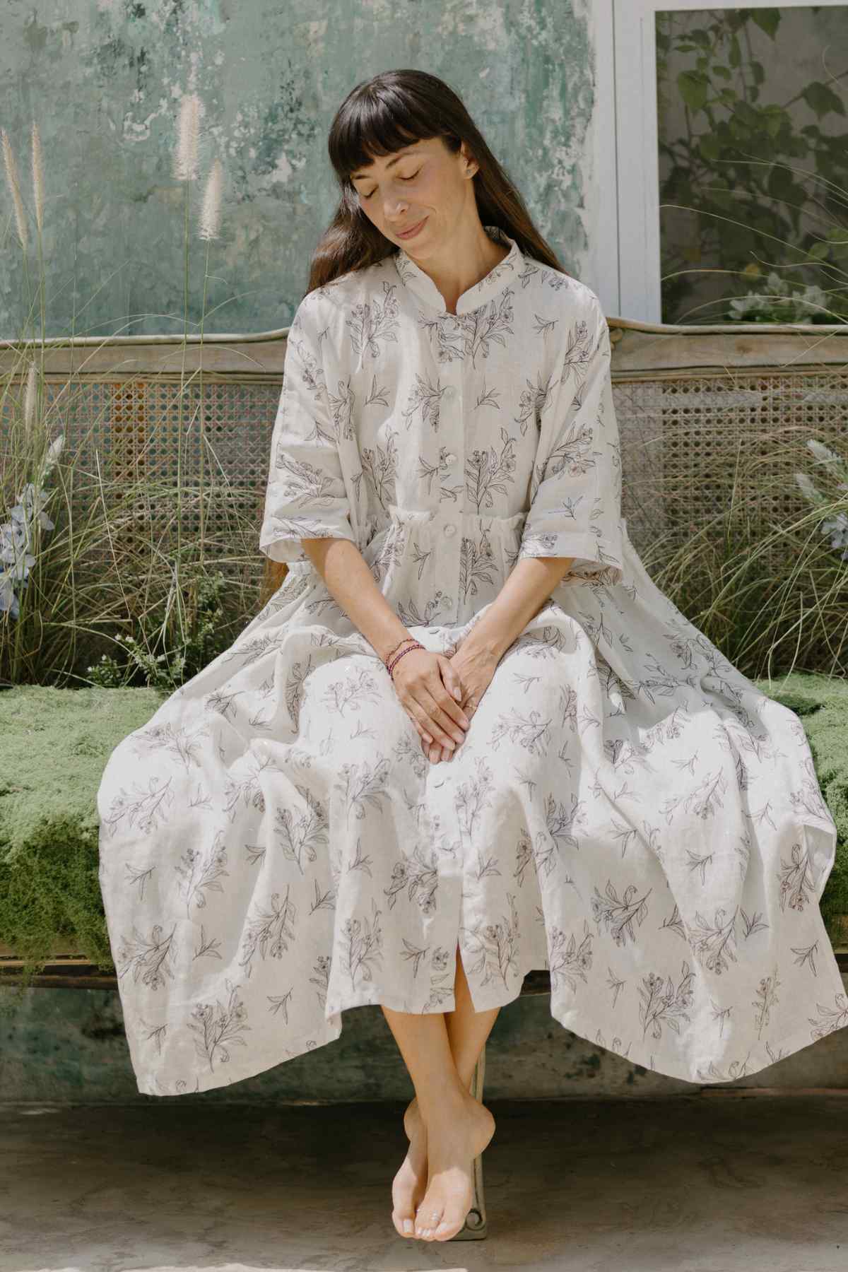 A person with long, dark hair sits barefoot on a moss-covered bench. They are wearing the limited edition Bona Dress from Myrah Penaloza's Cha Dao Tea Collection, featuring a beautiful floral pattern with a Tea Leaf Eco Print. The background showcases a rustic wall adorned in green and gray hues. Relaxed, their eyes are closed as they soak in the serene atmosphere.