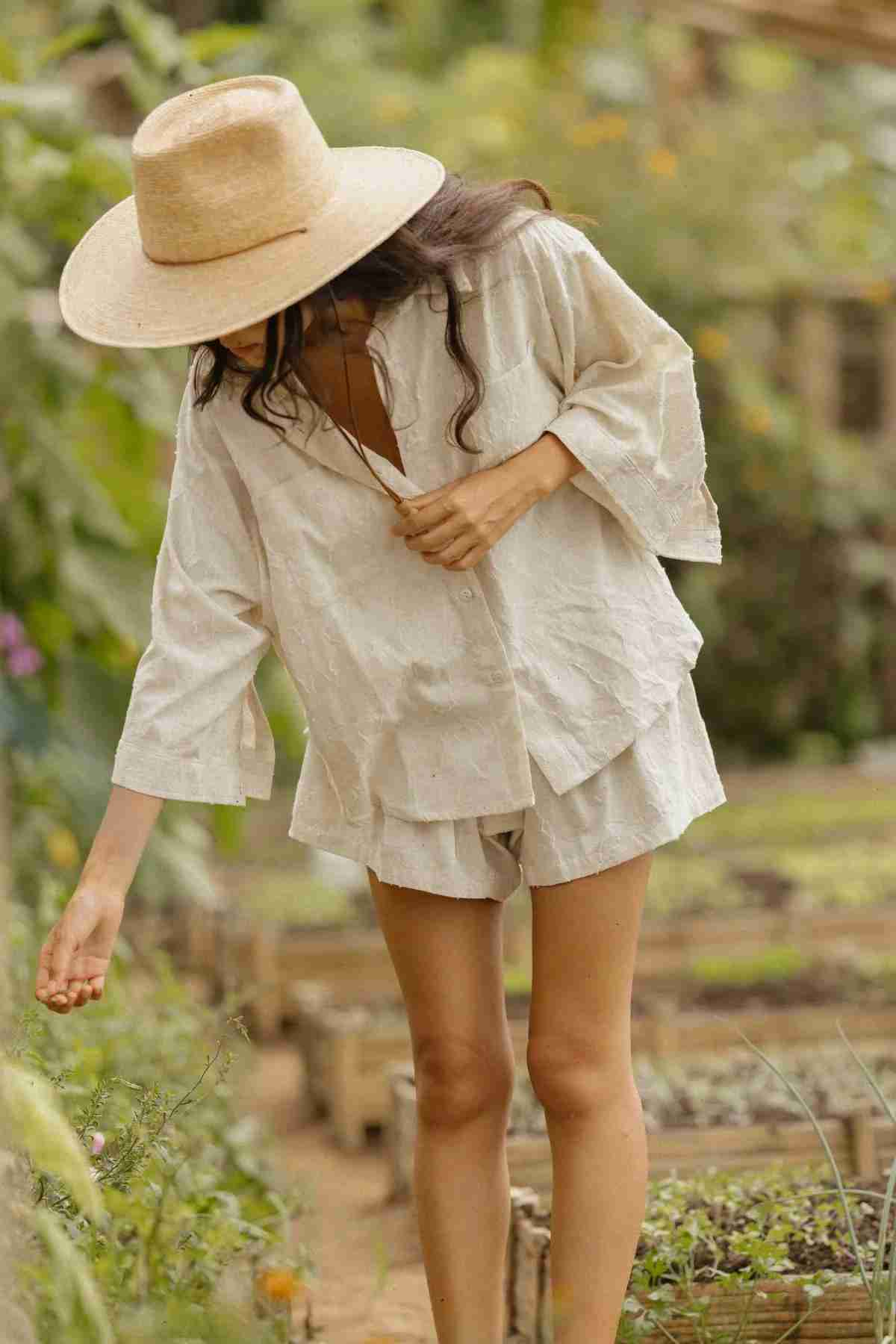 Dressed in the Linen Nidra Button Down Set from Myrah Penaloza and a straw hat, someone leans forward to tend to plants in a serene garden setting. Encircled by lush greenery and raised garden beds, this limited edition scene perfectly embodies the tranquility and gentle embrace of nature.