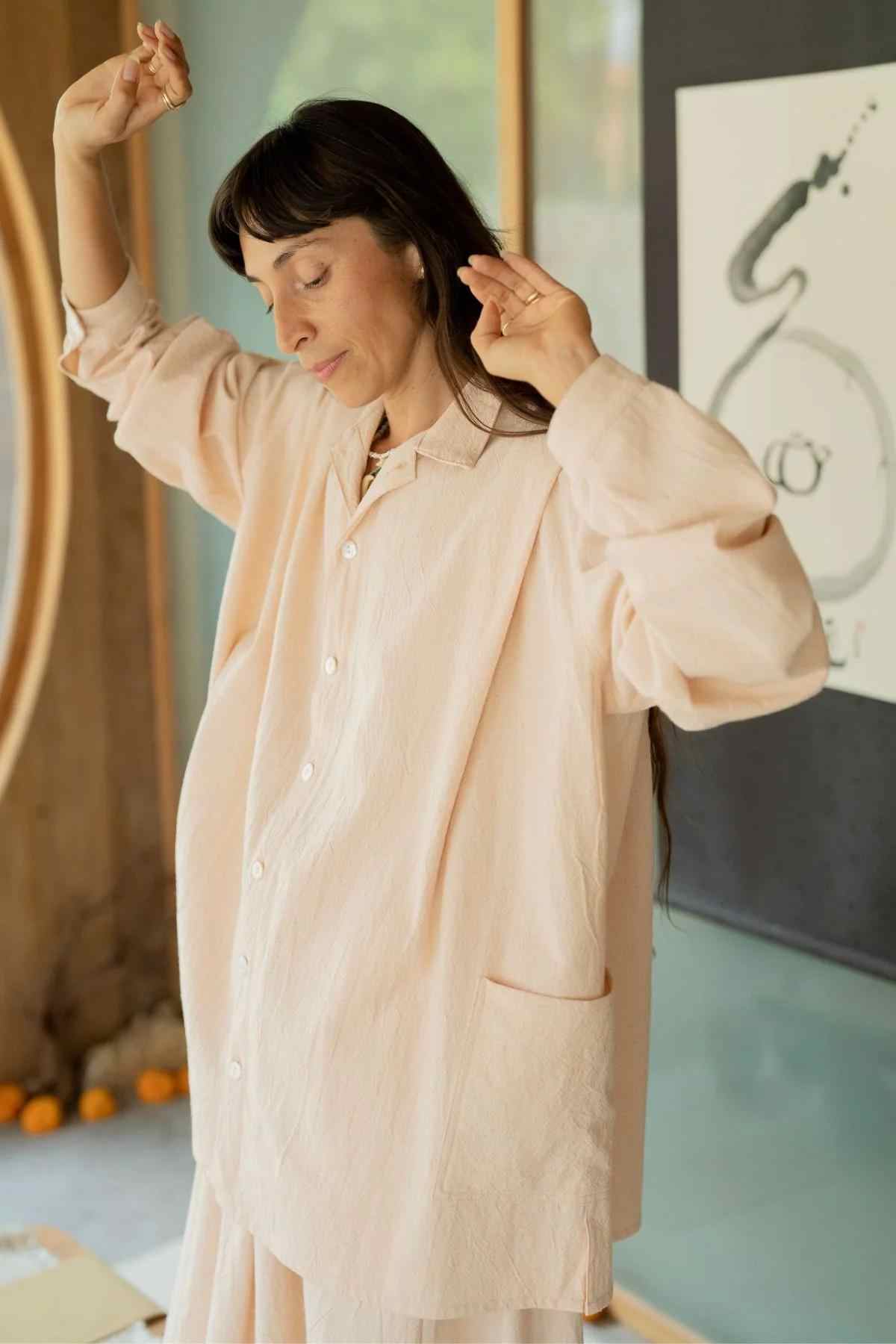 A person with long hair is standing with eyes closed, arms raised and slightly bent, fingers gently touching. They are wearing the Tender Jacket & Pant Set (3 of a kind) from Myrah Penaloza, featuring a loose, light-colored shirt paired with wide-legged pants. In the background, an abstract artwork adorns the wall and a window allows natural light to pour in.