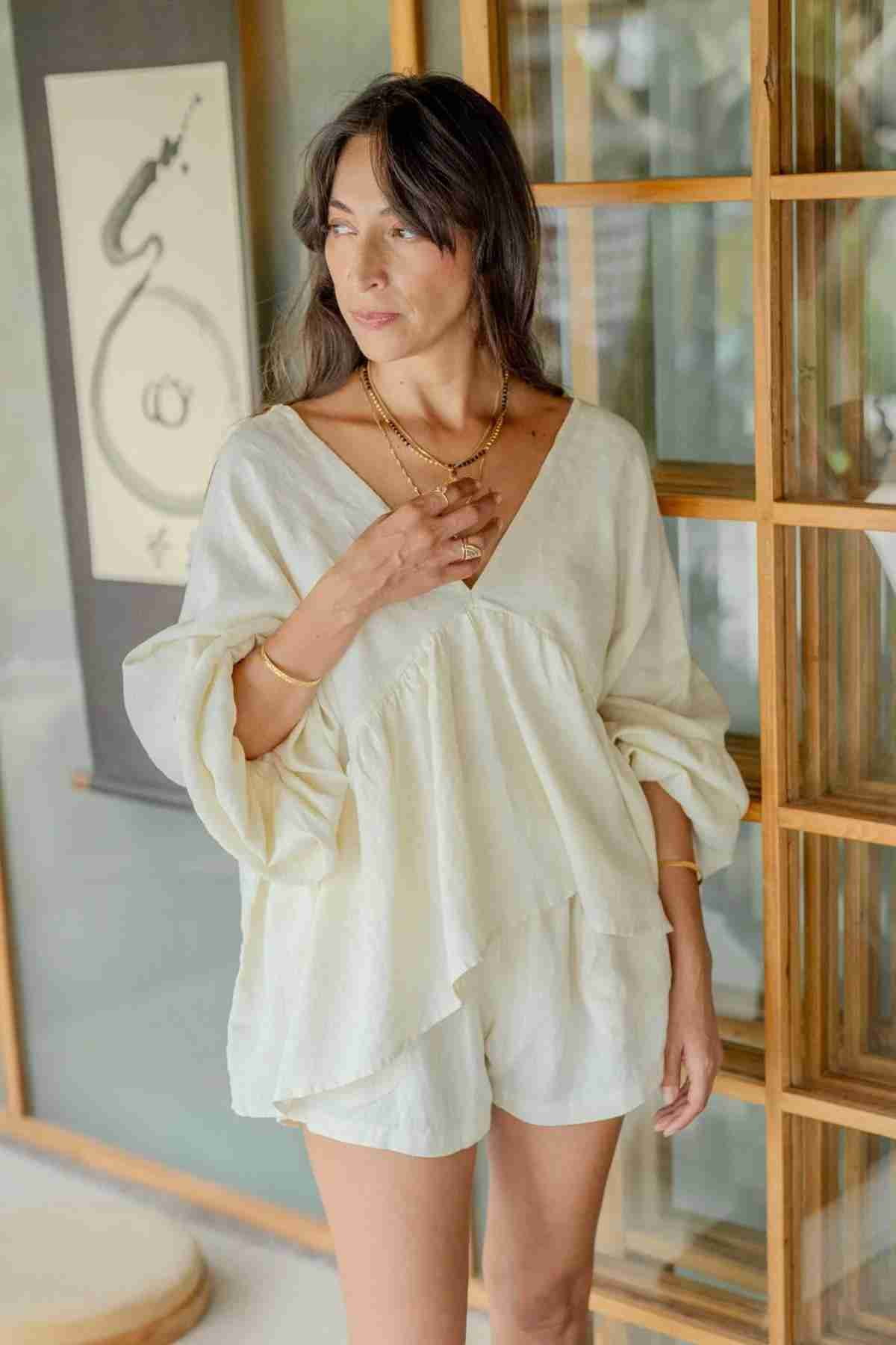 A woman stands in a room wearing an Eva Linen Top by Myrah Penaloza, complete with delicate flowering sleeves and matching shorts. She glances to the side, her hand gently resting near her chest. Behind her is a wooden-framed glass door and wall art featuring a snake-like design.