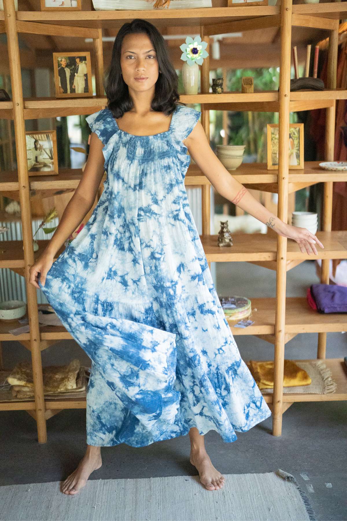 A person wearing the Anna Yemaya Hand Tye Dyed Indigo gown by Myrah Penaloza stands barefoot in front of a wooden shelf decorated with books and other ornaments, with a flower visible in the background. They gently extend the blue and white tie-dyed indigo dress with one hand.