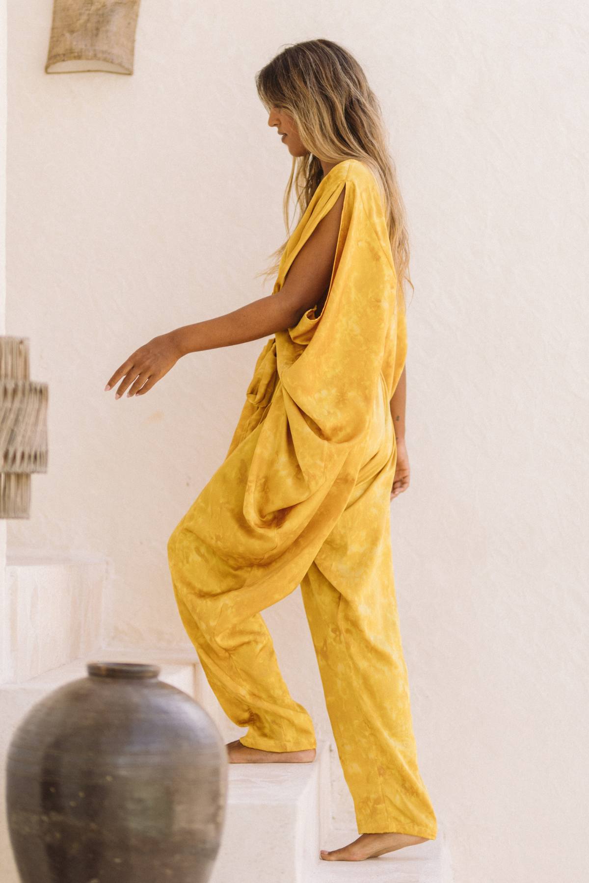 A person with long hair, dressed in a loose-fitting Kuan Yin Playsuit made from 100% silk and botanically dyed in Turmeric Gold by Myrah Penaloza, is walking up white stairs. A large, dark pot is visible at the bottom of the stairs, and the background features a white, textured wall. The scene appears to be bright and airy.