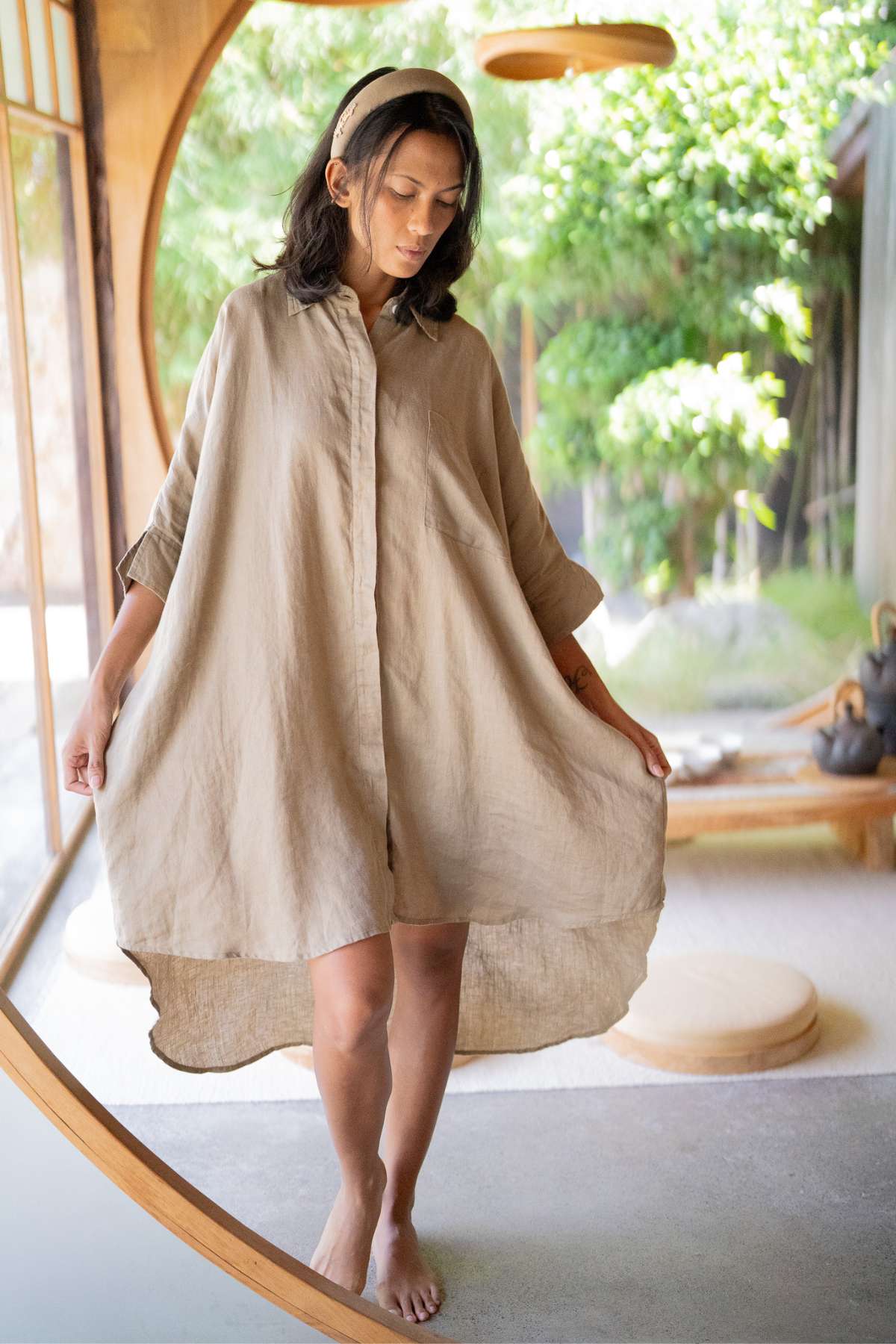 In a tranquil setting with expansive circular windows, a person stands barefoot in the serene room, adorned in the Linen Suka Button Down Long Top - BF from Myrah Penaloza. The soft beige hue of the dress harmonizes with the gentle lighting and glimpses of verdant greenery outside. This scene beautifully showcases versatile styling potential while highlighting eco-friendly fabric choices.