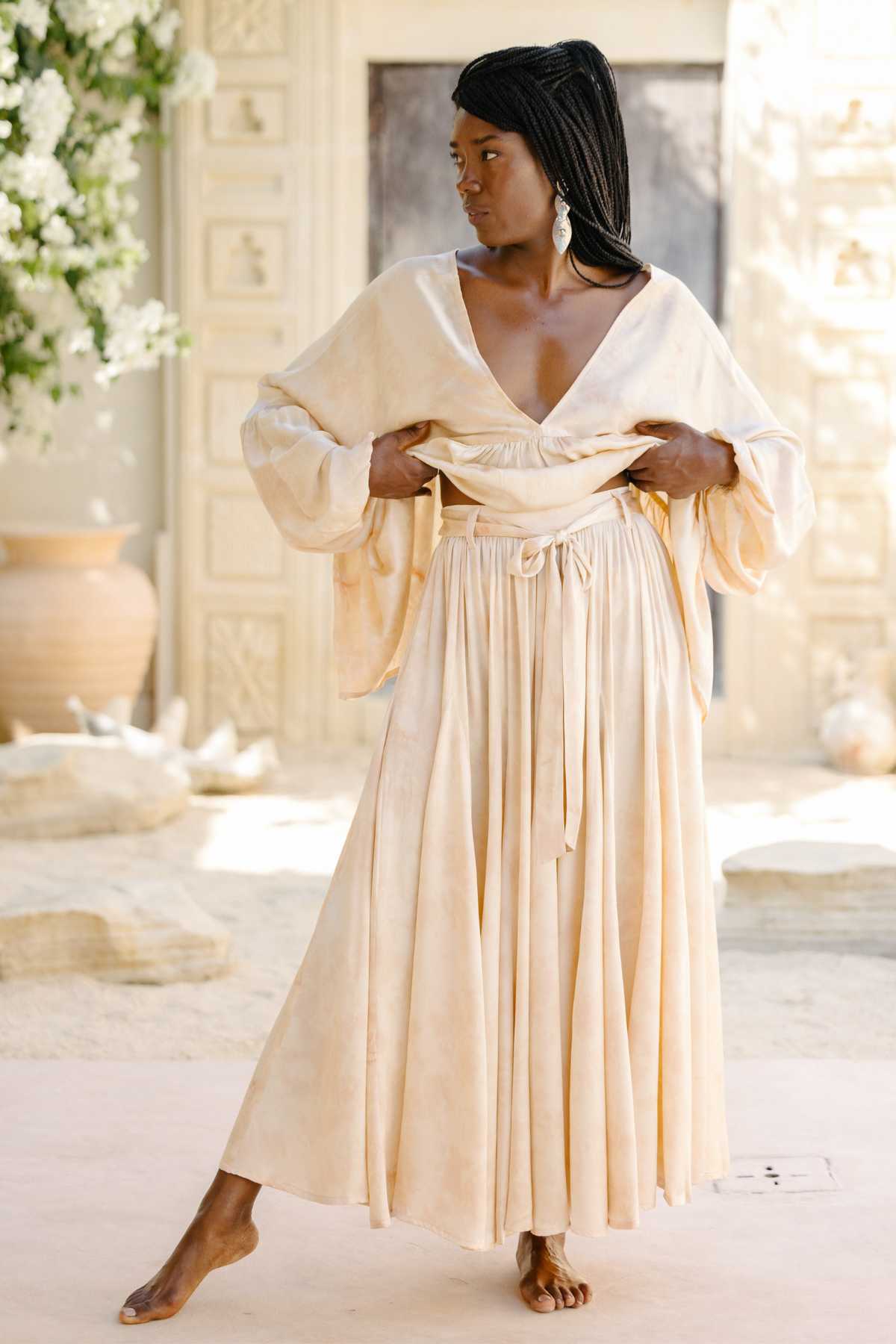 A woman stands in a sunlit room wearing the elegantly flowing Eva Top & Orisa Dance Skirt Set, botanically hand-dyed in silk by Myrah Penaloza. She adjusts the top with one hand. The background features a large vase, stone textures, and soft floral decor.