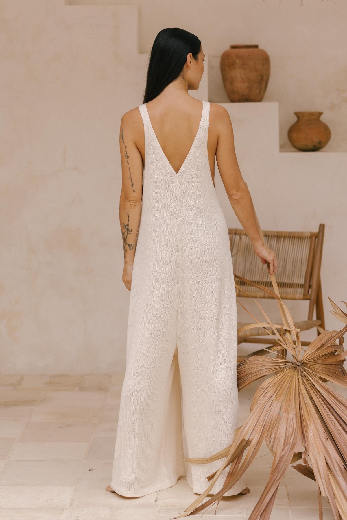 A woman stands with her back to the camera, wearing the Myrah Penaloza Kundalini Playsuit, a limited edition piece crafted from cream-colored cotton knit. The playsuit features a gracefully sleeveless design and a plunging back. Her long dark hair cascades over her tattooed arms in a setting adorned with neutral-toned decor, such as ceramic pots and a wicker chair.