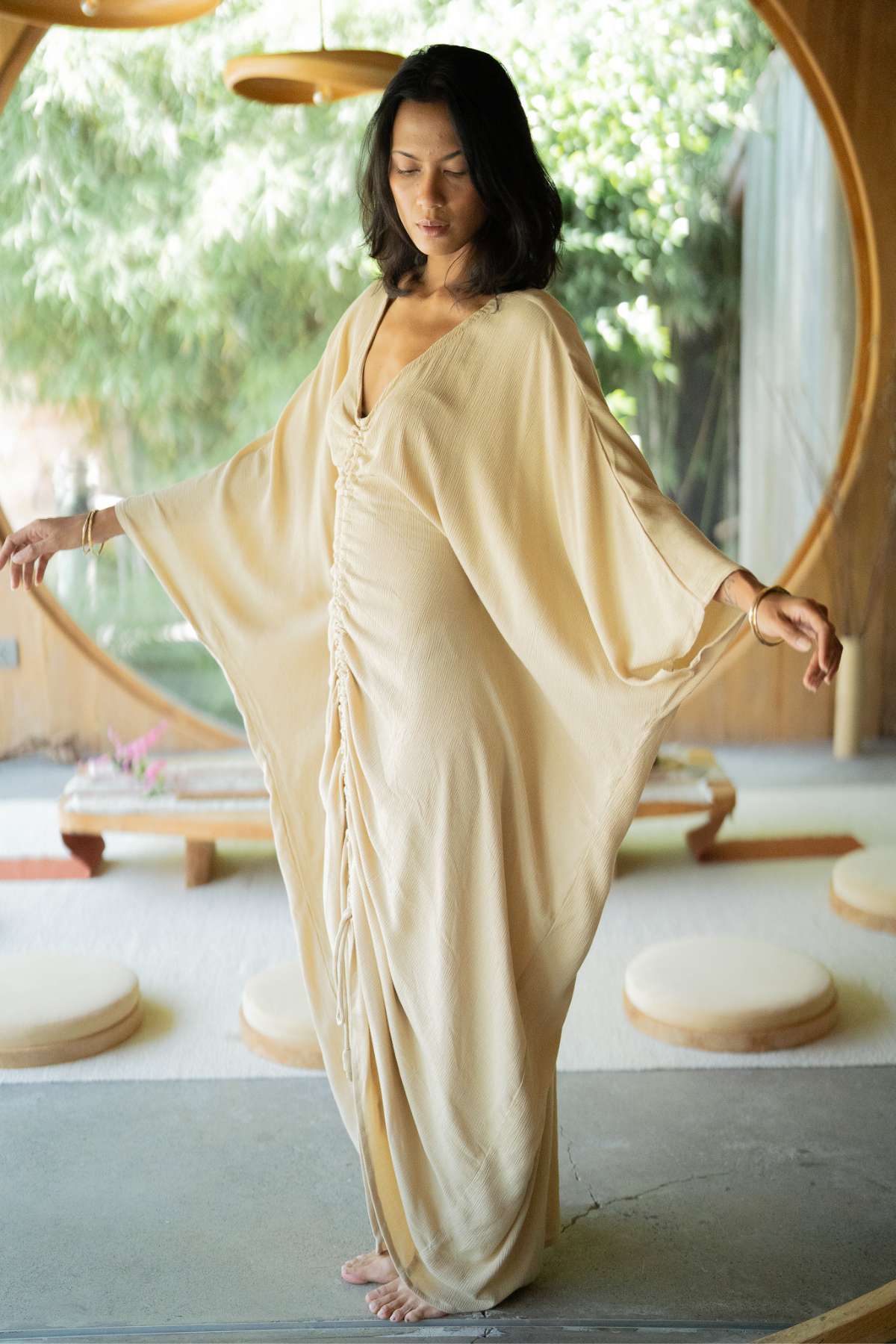 In a tranquil room filled with sunlight streaming through a circular window, a woman stands barefoot, wearing the flowing Virgo Moon kaftan by Myrah Penaloza. The long sleeves and intricate detailing of her attire are beautifully illuminated as she gracefully extends her arms, exuding a sense of calm.